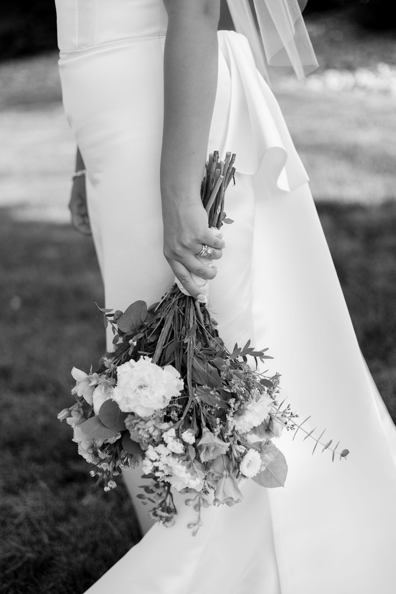 Black and white photo of a bride's hands holding a delicate bouquet with white flowers and greenery against her dress.