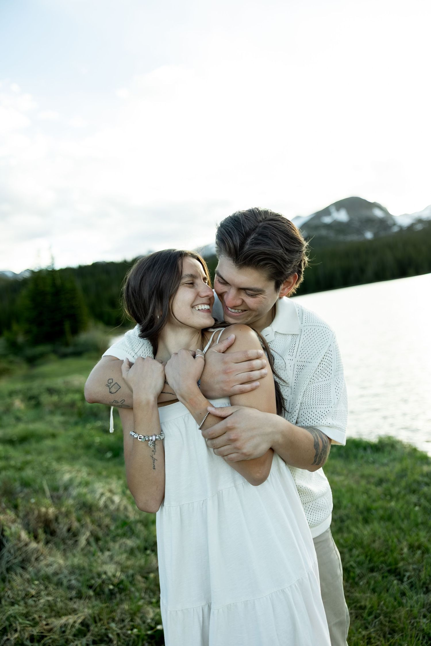 A couple embraces near a mountain lake, the man hugging the woman in a white dress from behind as she smiles joyfully.