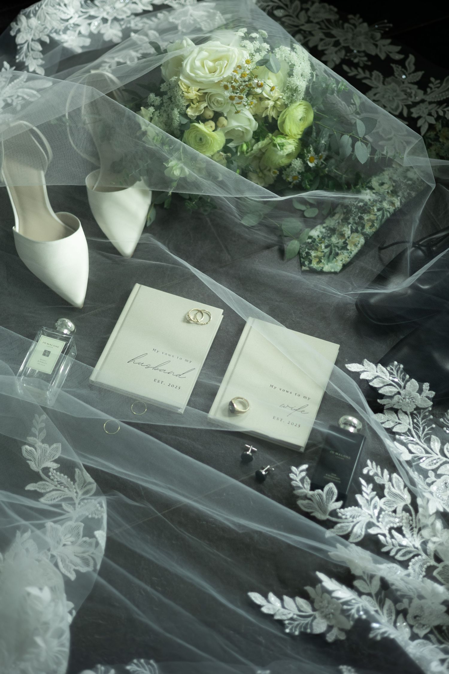 Elegant wedding flat lay with lace veil, white heels, floral bouquet, gold rings, cufflinks, and stationery on dark background.