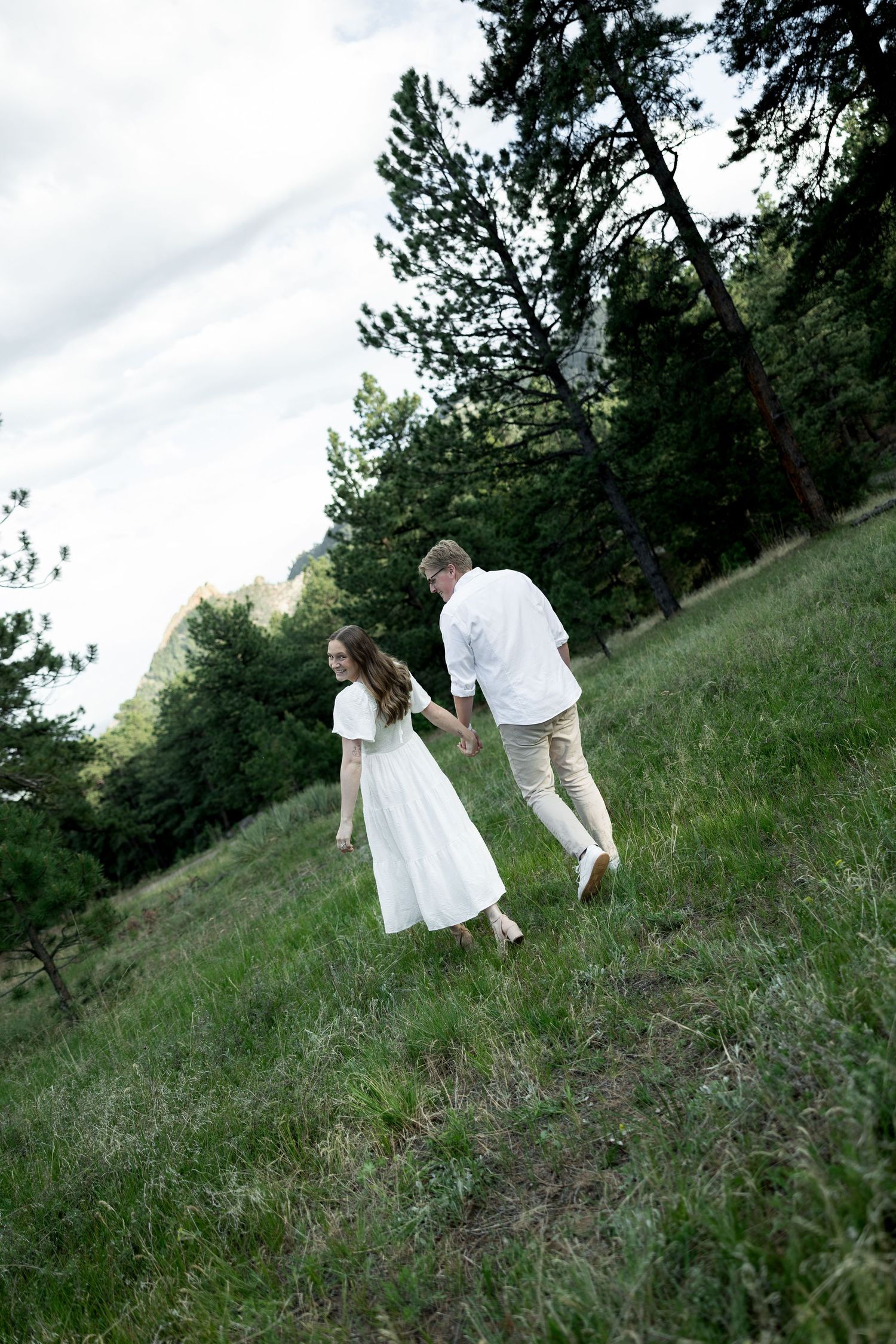 Couple holding hands walking through a lush green meadow surrounded by pine trees and rocky mountains.