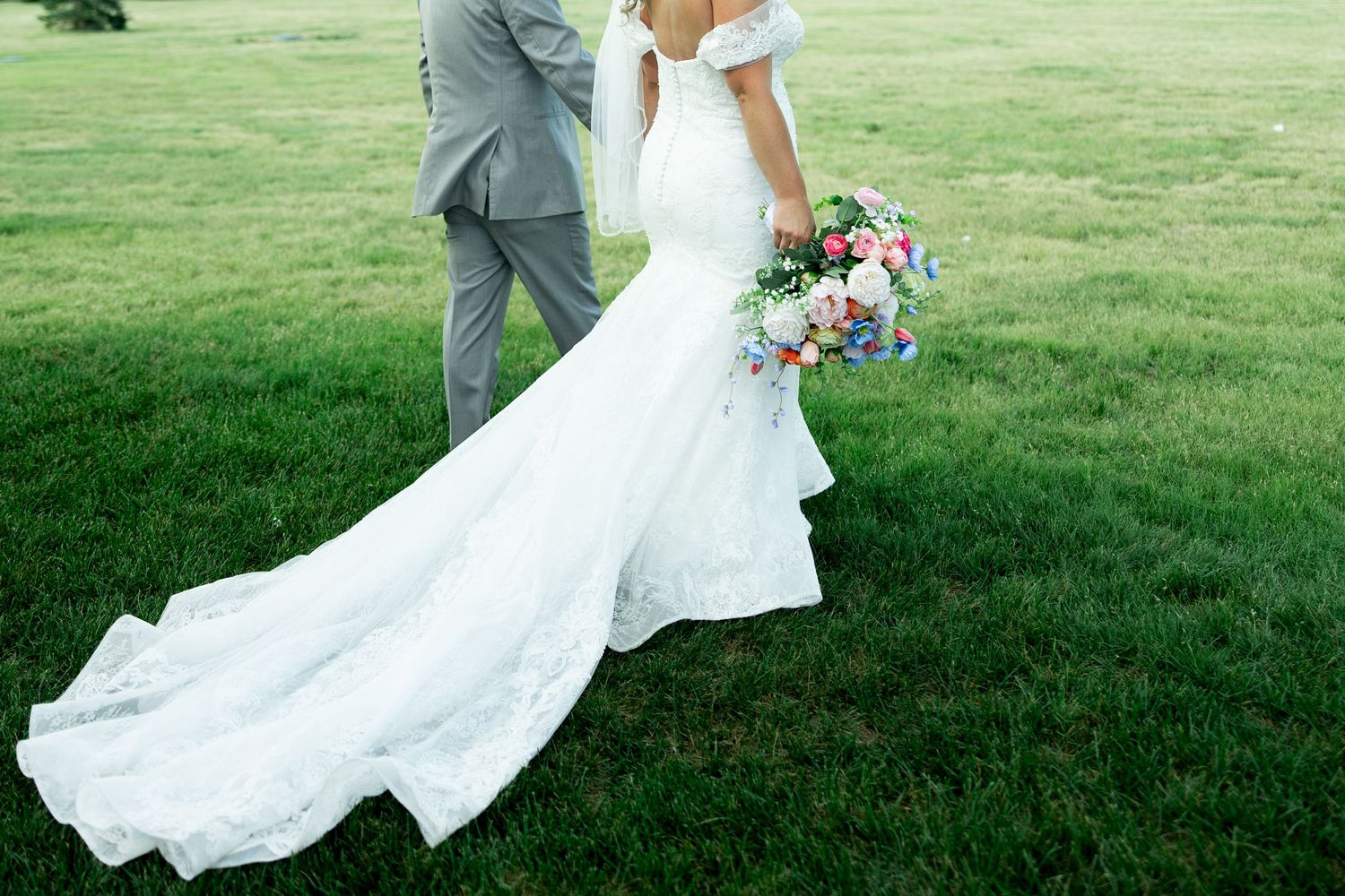 Bride in white lace mermaid gown with long train holds colorful bouquet beside groom in gray suit on green lawn.
