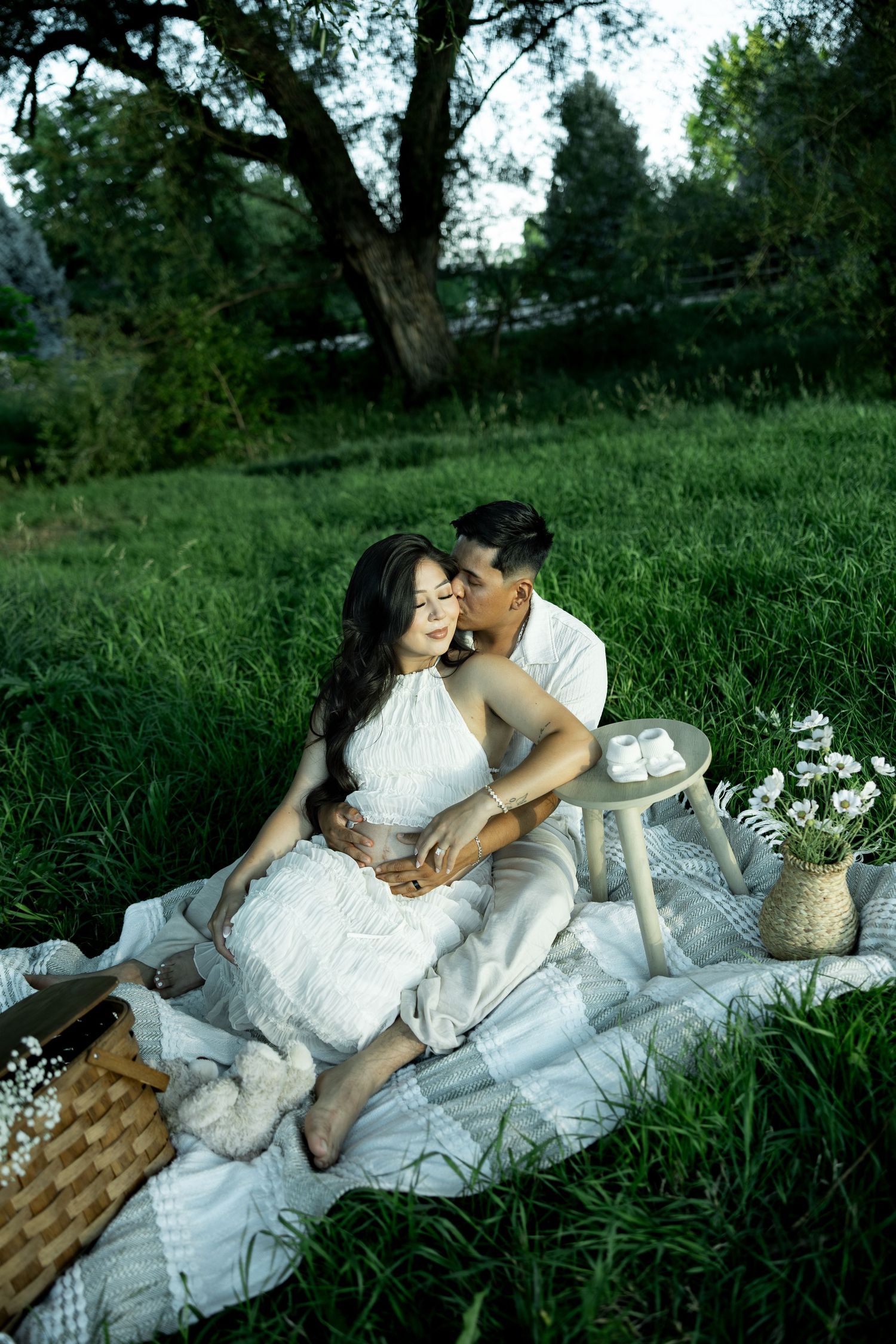 Couple sharing a romantic kiss on a white picnic blanket in a lush green park, with a basket and flowers nearby.