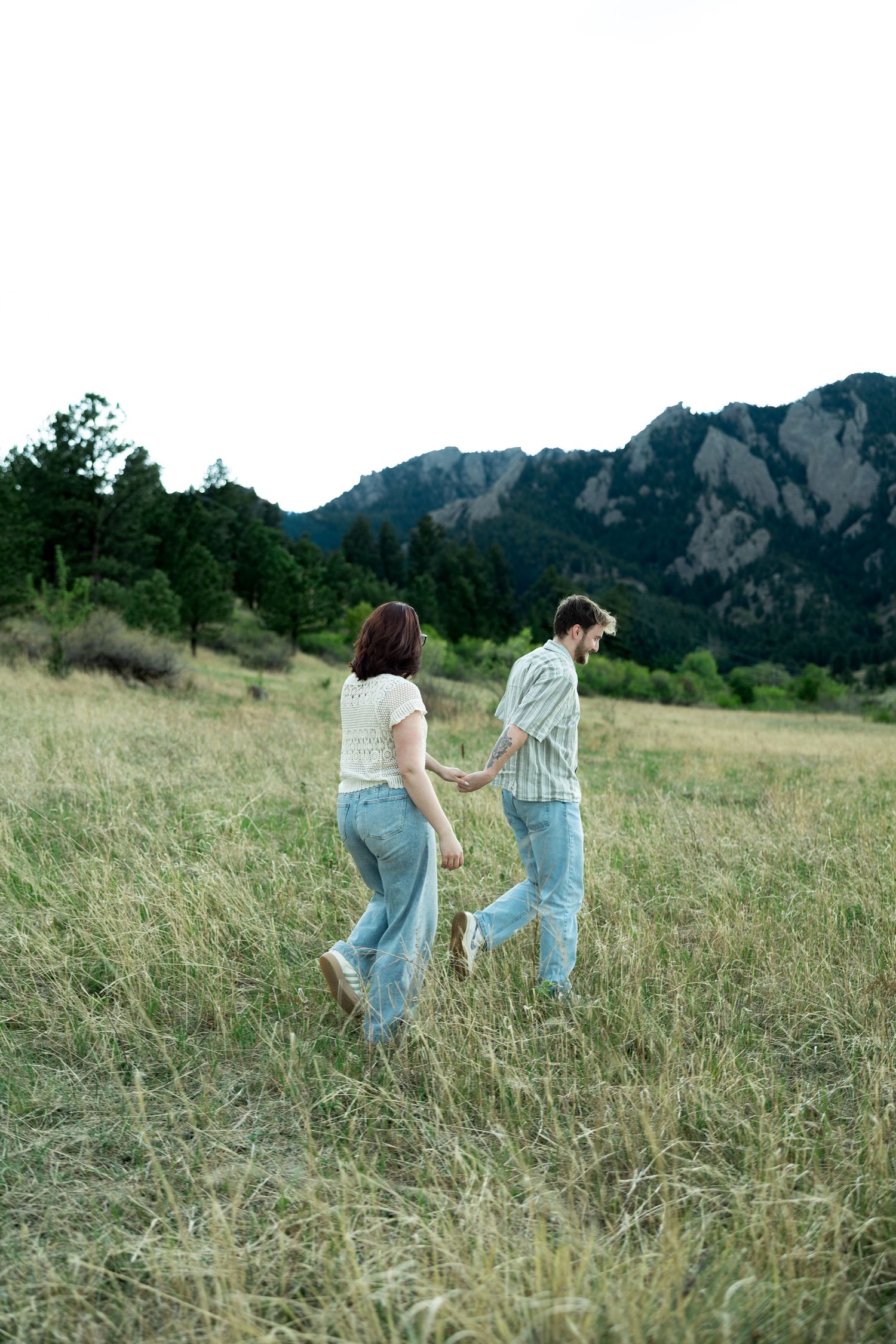 A couple holds hands while walking through a grassy mountain meadow, both wearing jeans and casual tops.