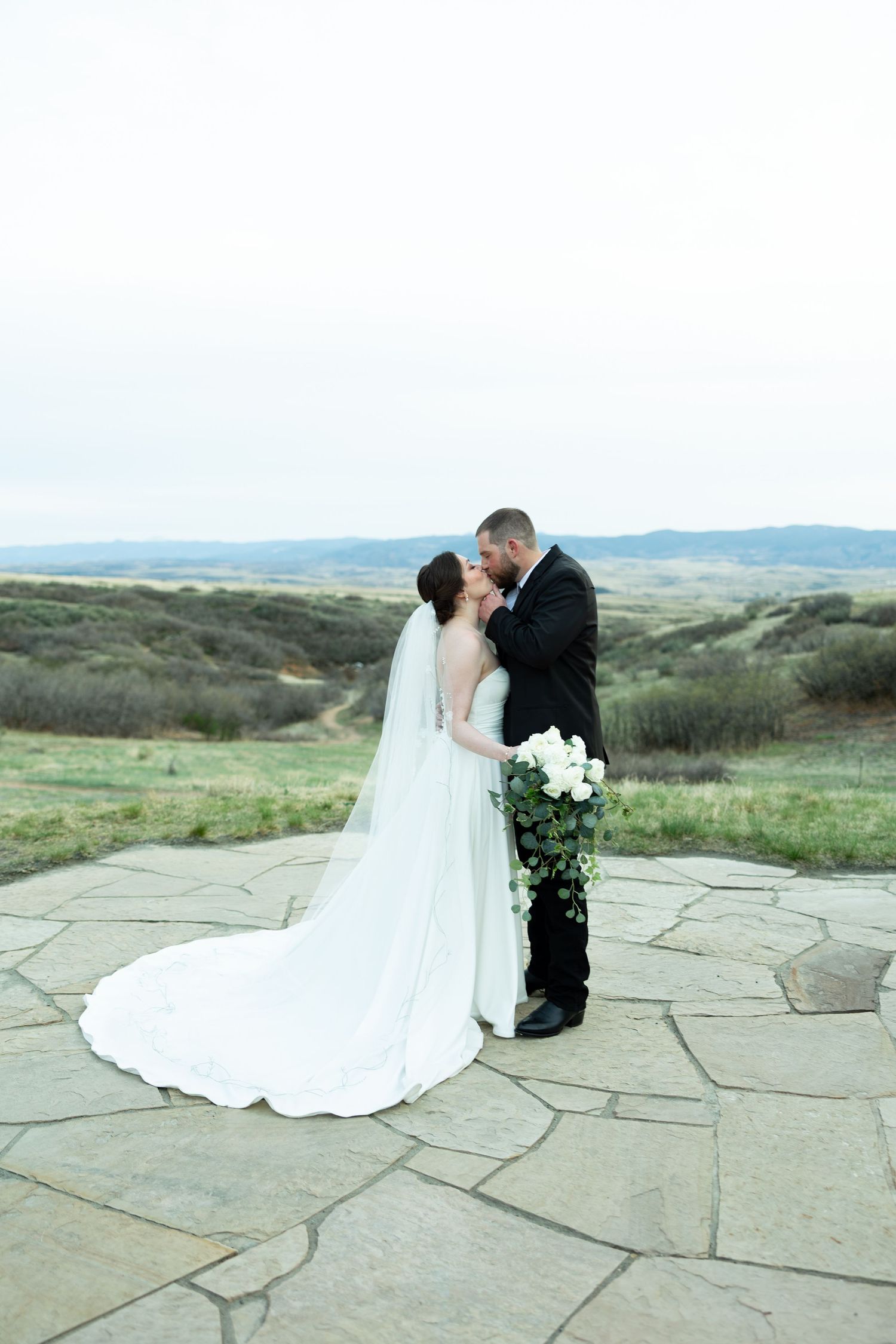 Bride and groom share a romantic kiss on a stone patio with scenic rolling hills backdrop on their wedding day.