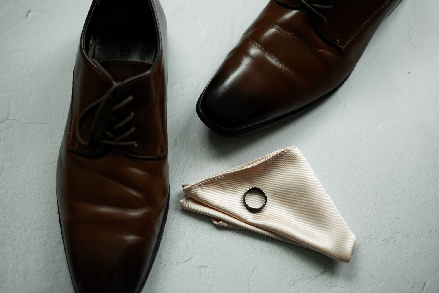 Brown leather dress shoes with beige pocket square and wedding ring on light gray surface.