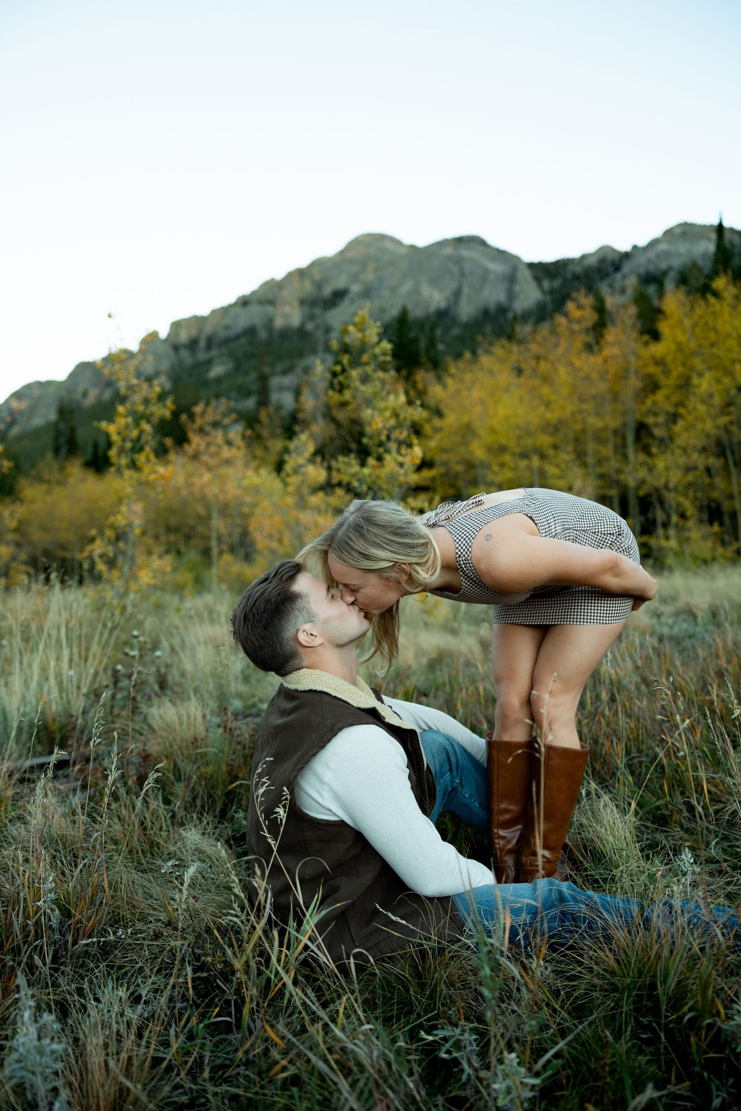 Couple kissing in a grassy mountain meadow, the woman in brown boots leaning down to the seated man, with golden aspen trees behind them.