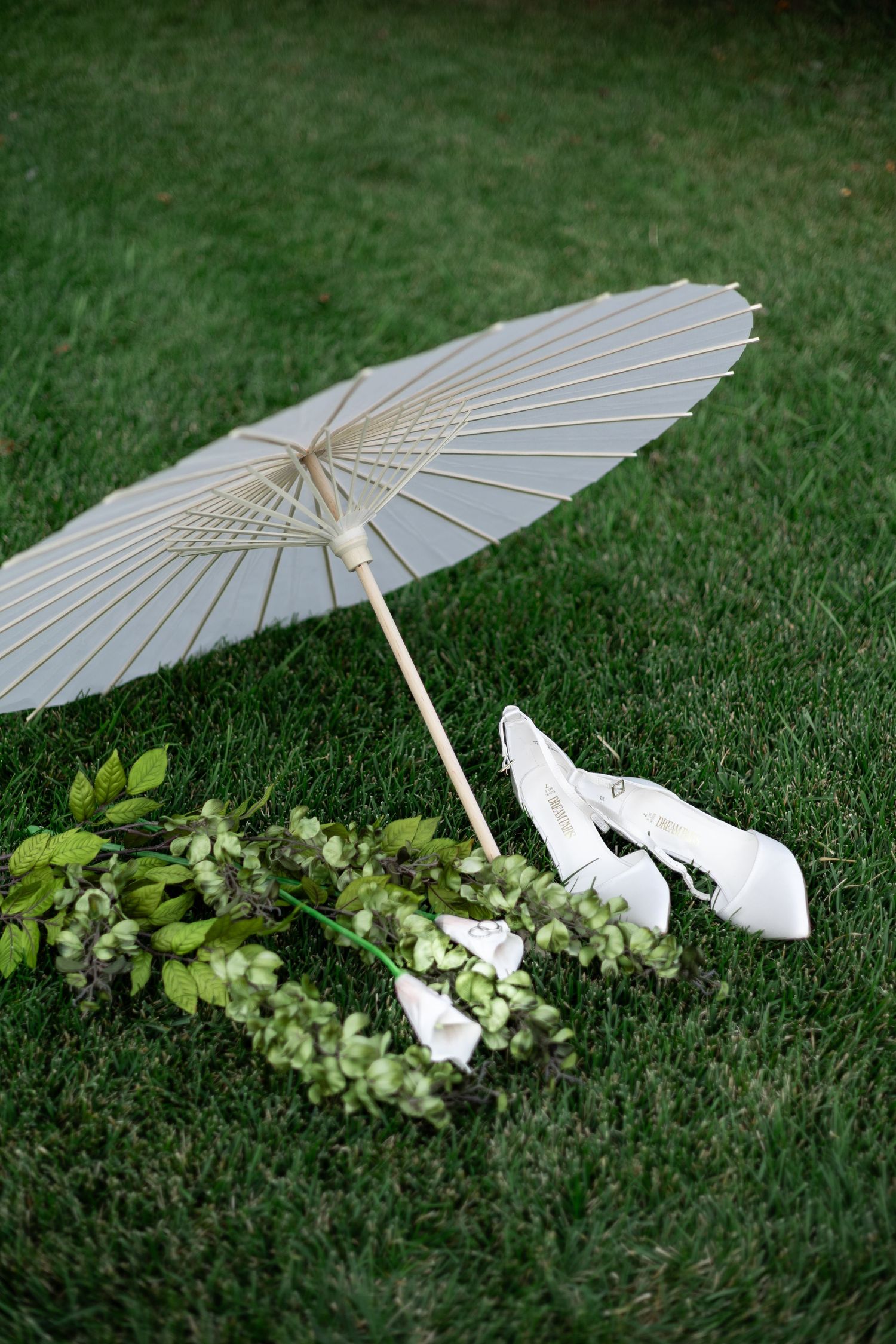 White paper parasol and elegant gloves rest on lush green grass next to delicate foliage in an outdoor setting.