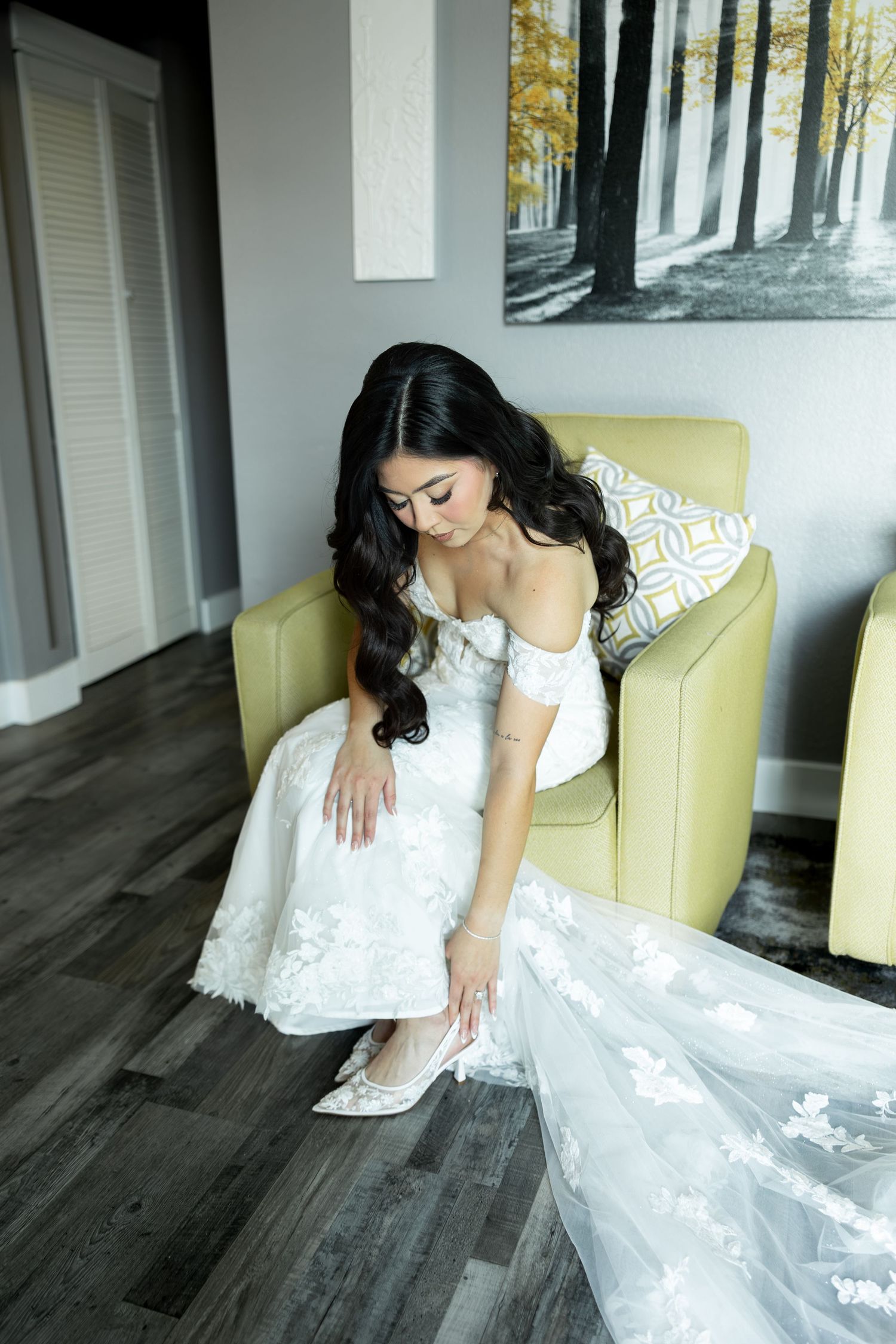 A bride in a white lace wedding dress sits elegantly on a yellow chair against a grey wall.