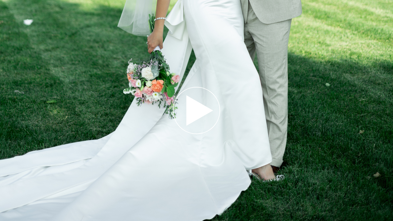 Bride in white gown holding colorful bouquet stands with groom in tan suit on green grass at outdoor wedding.