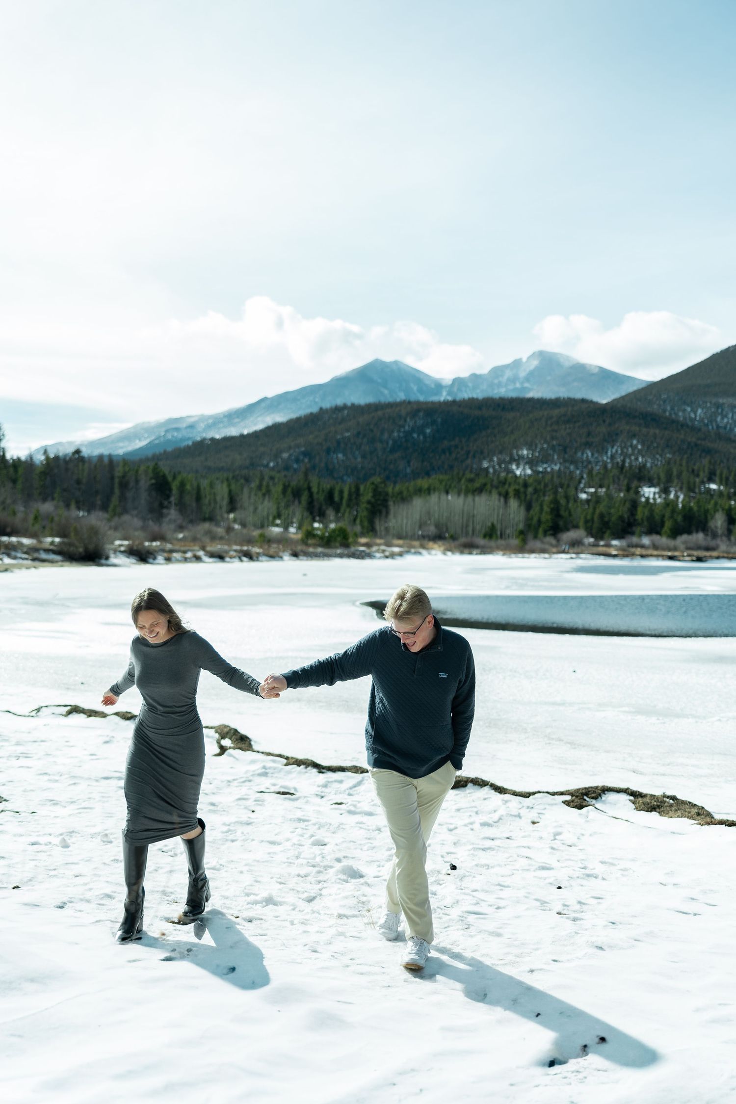 Couple holding hands walking on a snowy frozen lake with mountain backdrop in winter landscape.