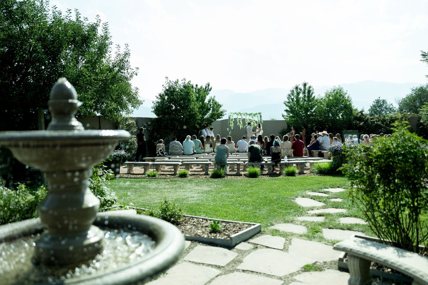 Outdoor garden wedding ceremony with guests seated near a stone fountain and lush greenery on a sunny day.
