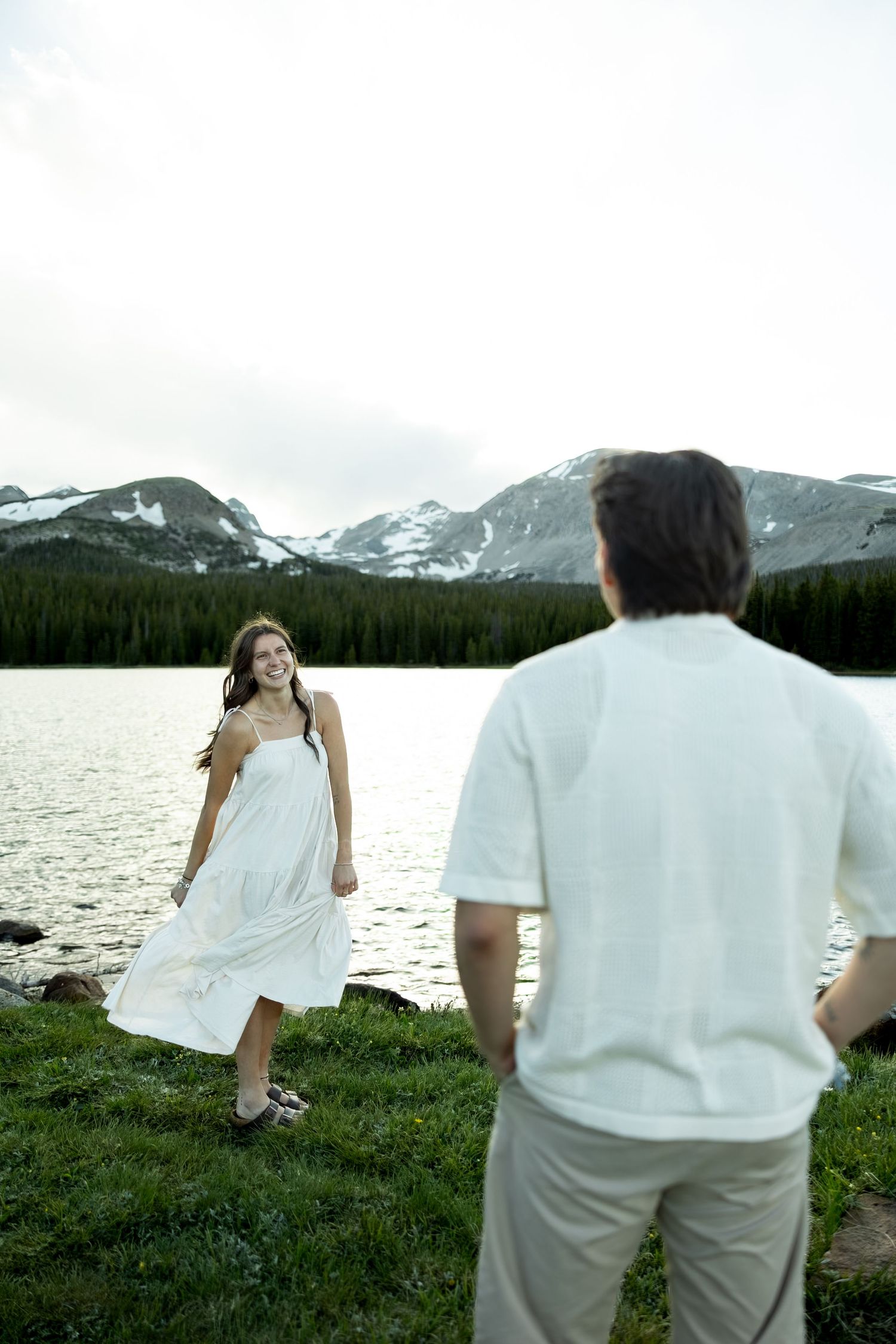 Smiling woman in white dress stands by mountain lake as man in white shirt watches her, with snow-capped peaks in background.