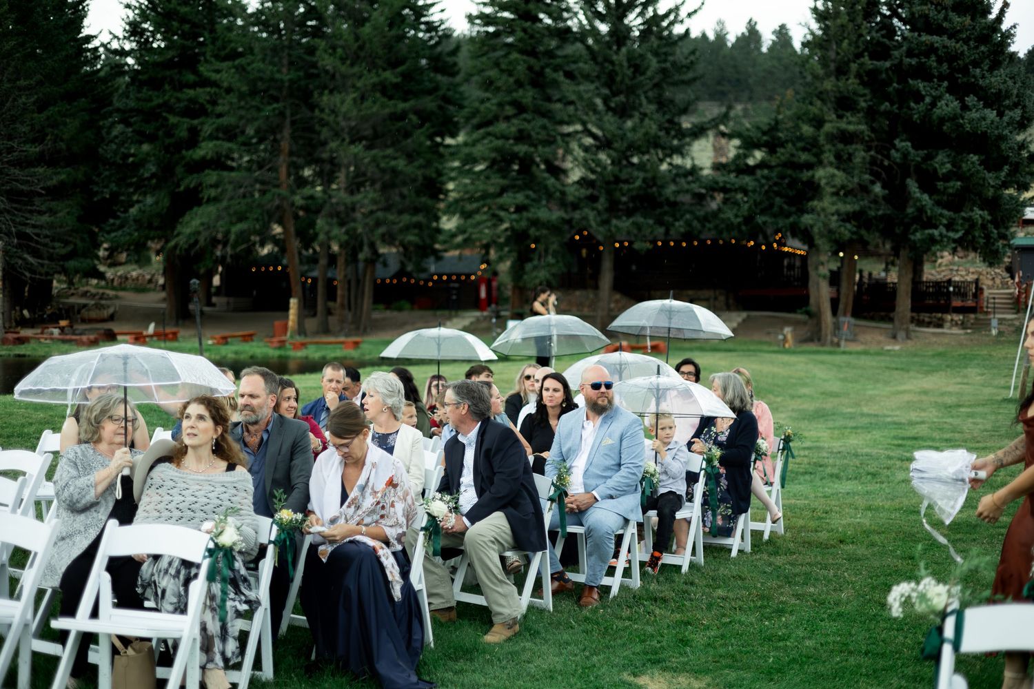 Outdoor wedding ceremony setup with white chairs and guests holding umbrellas in a wooded setting.