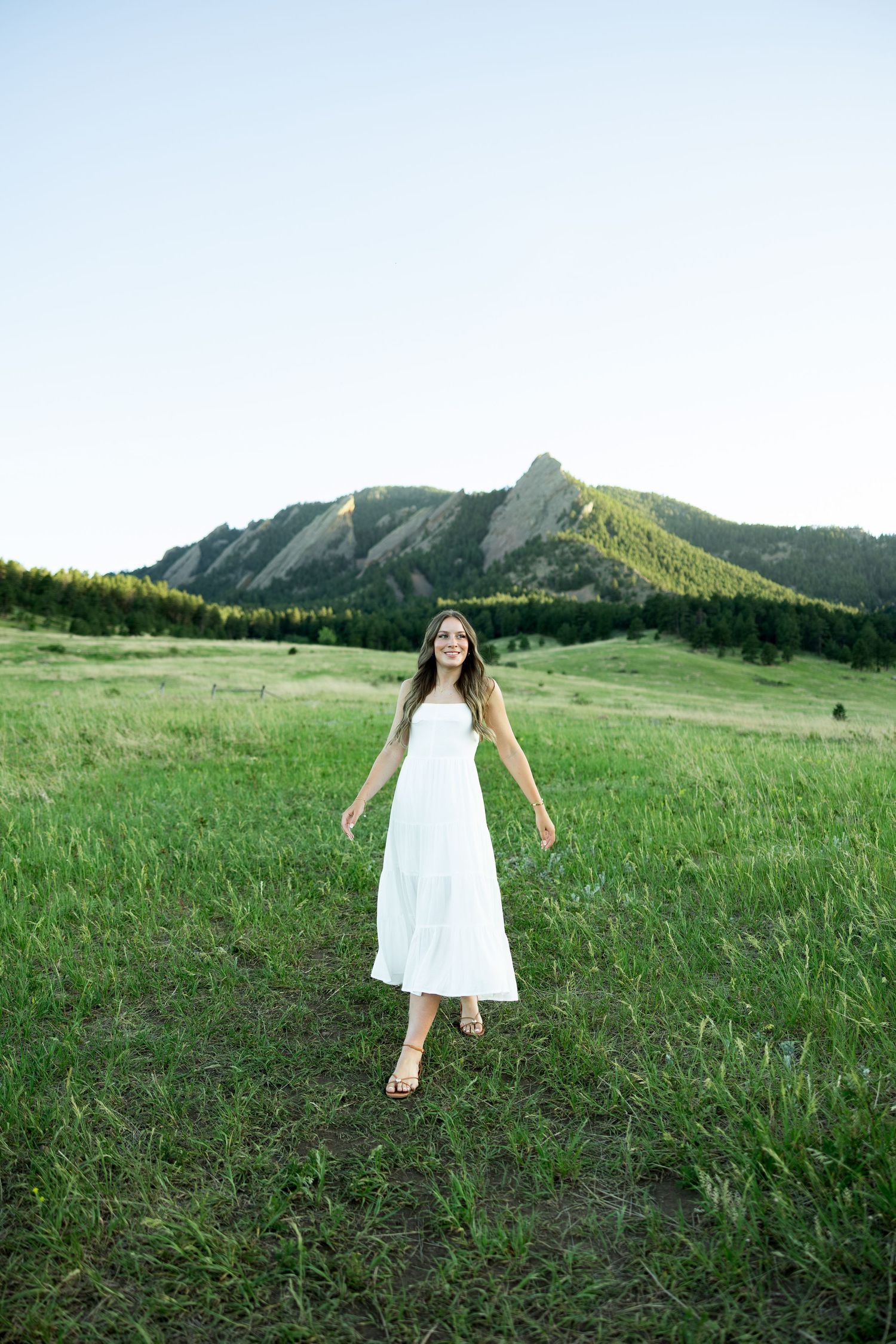 Woman in white sundress walking through green meadow with rocky Flatirons mountain backdrop.