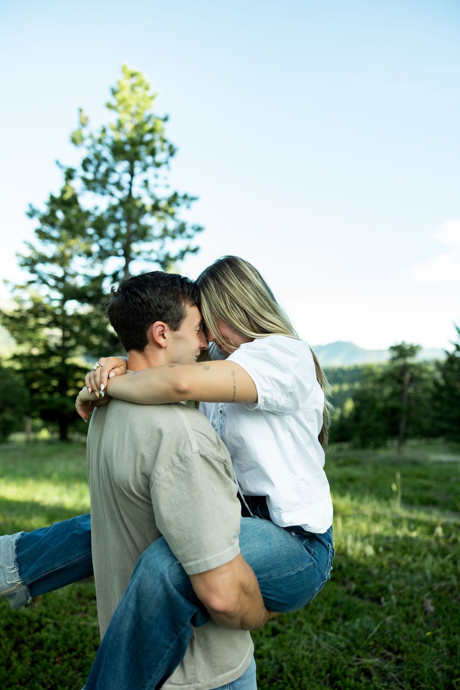 A man lifts and embraces a woman outdoors near pine trees, sharing a romantic moment in a lush green mountain setting.