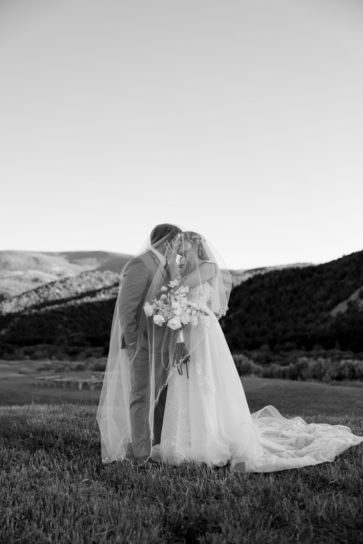 Black and white photo of bride and groom kissing under veil in mountain meadow landscape.