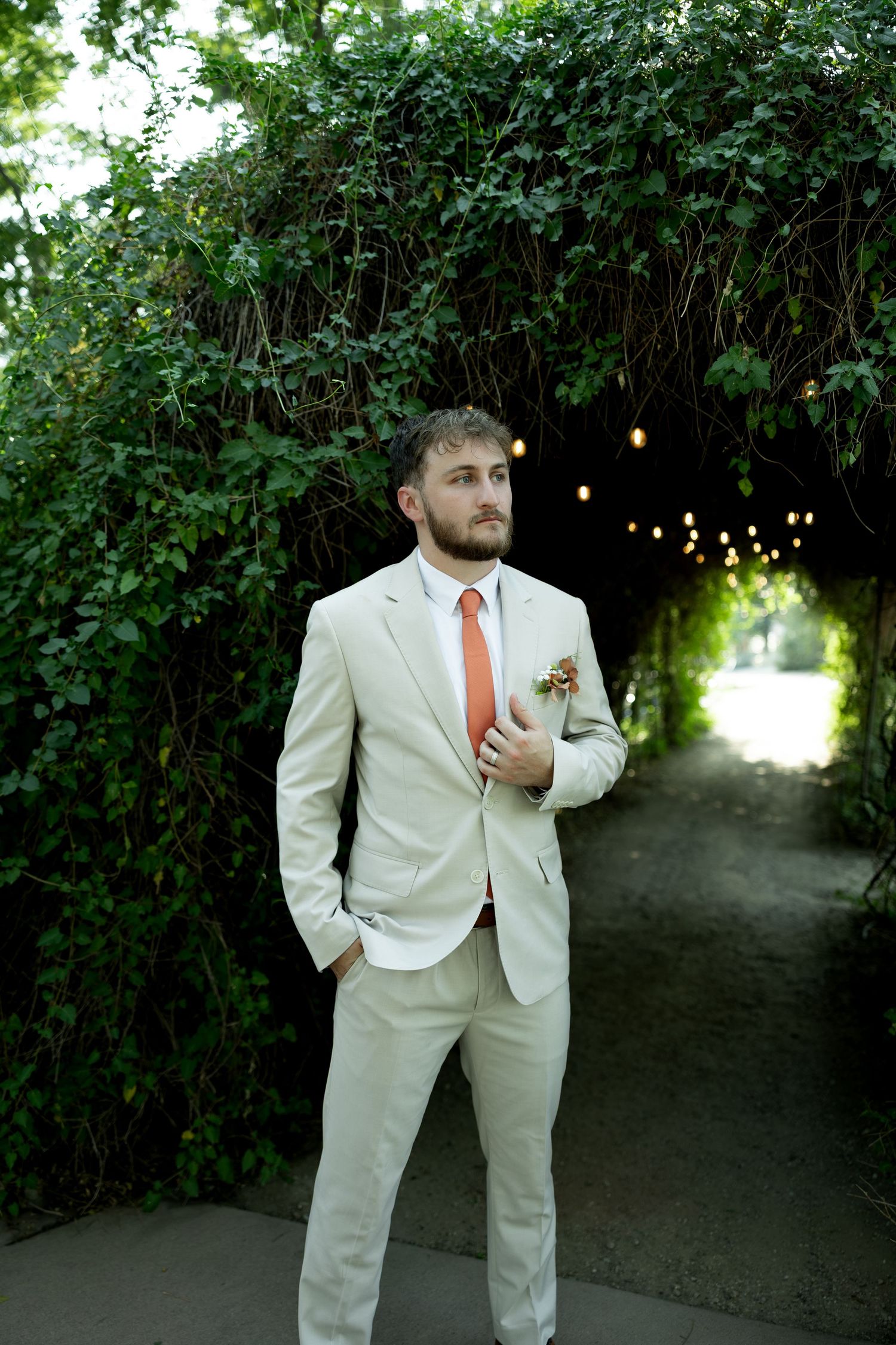 Groom in beige suit with orange tie stands under lush ivy arch with string lights at outdoor venue.
