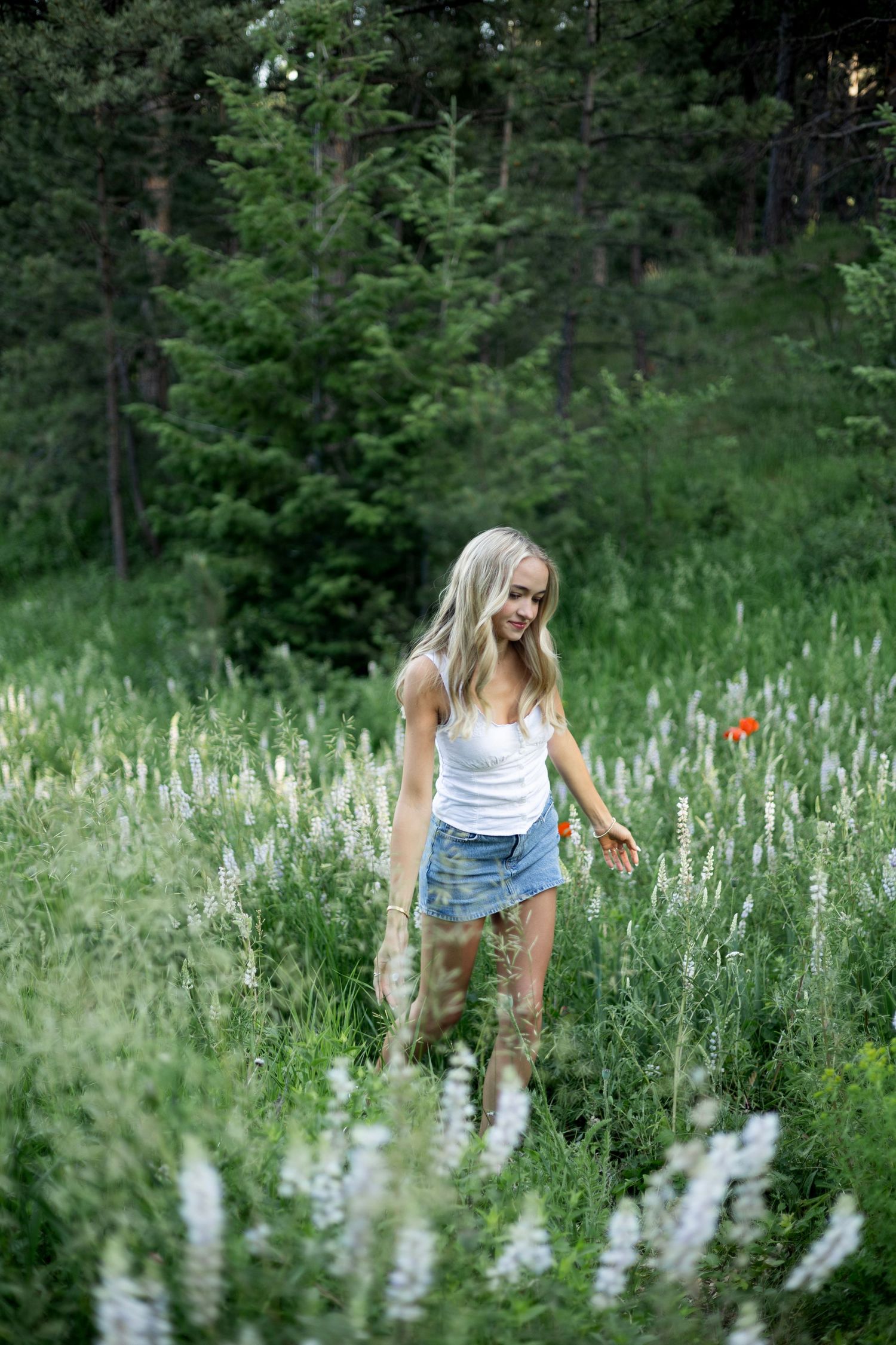 A person in denim shorts stands among blooming wildflowers and tall grass in a forest clearing.
