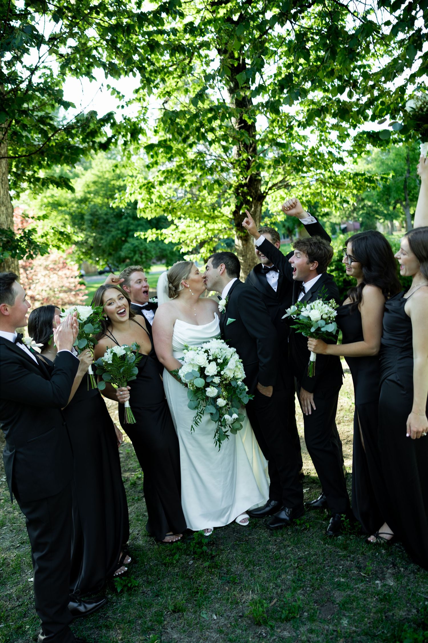 A joyful wedding party in black attire celebrates as the bride and groom share a kiss outdoors under green trees.