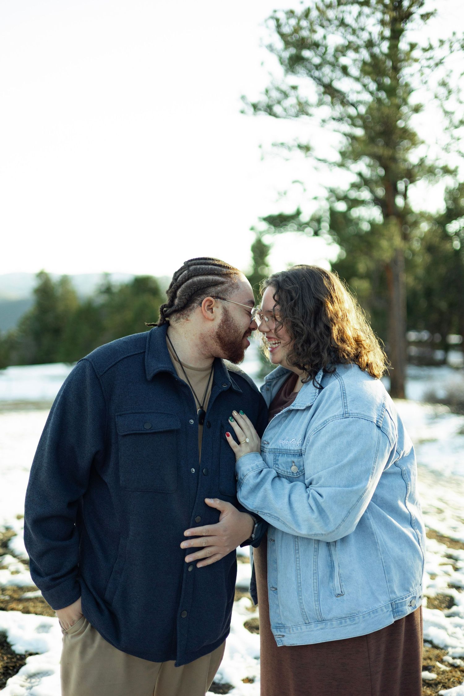 Couple sharing a romantic moment in a snowy winter landscape, wearing casual jackets among pine trees.