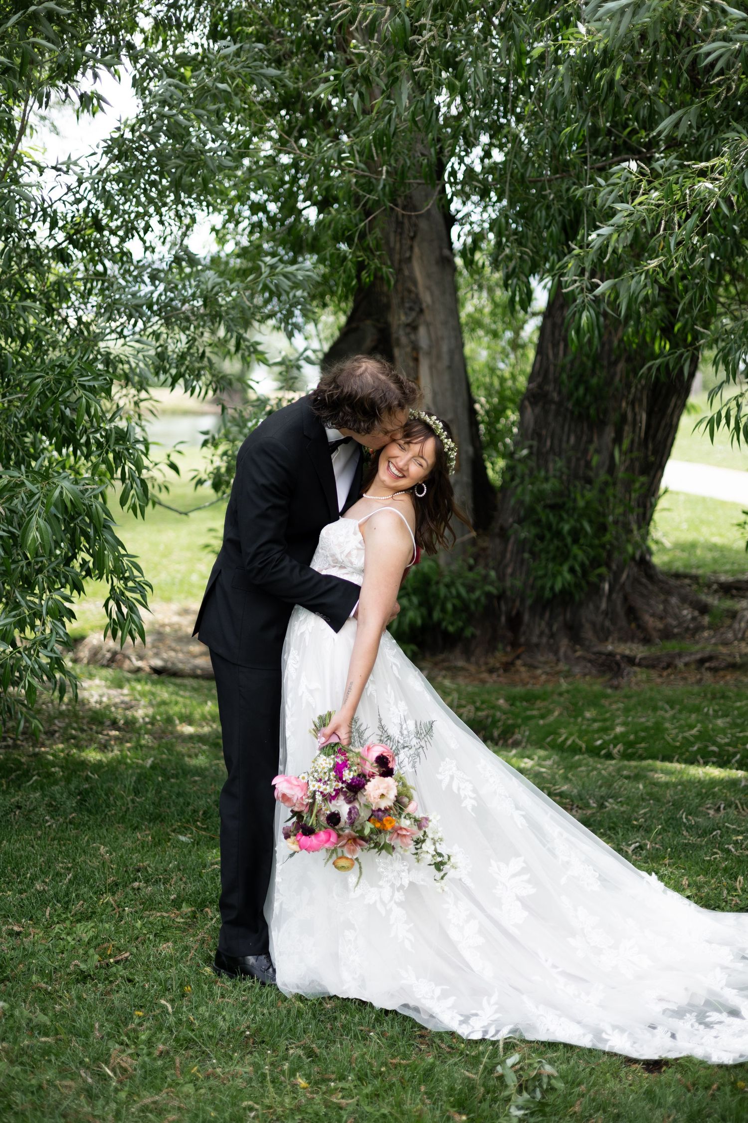 Romantic wedding couple embraces under willow tree in garden setting.