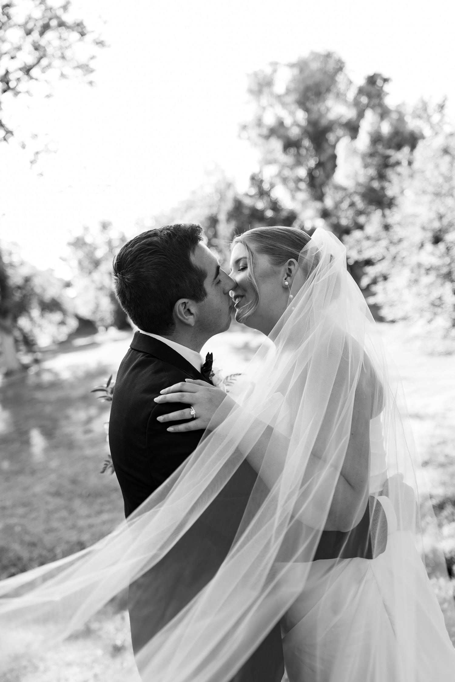 Black and white photo of a bride and groom sharing a romantic moment, her veil flowing, surrounded by trees.