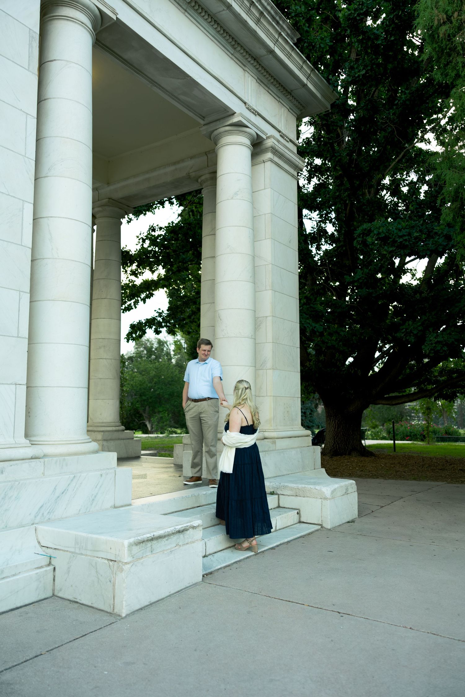 Couple on marble steps of classical building with columns and large tree, woman in navy dress, man in casual attire.
