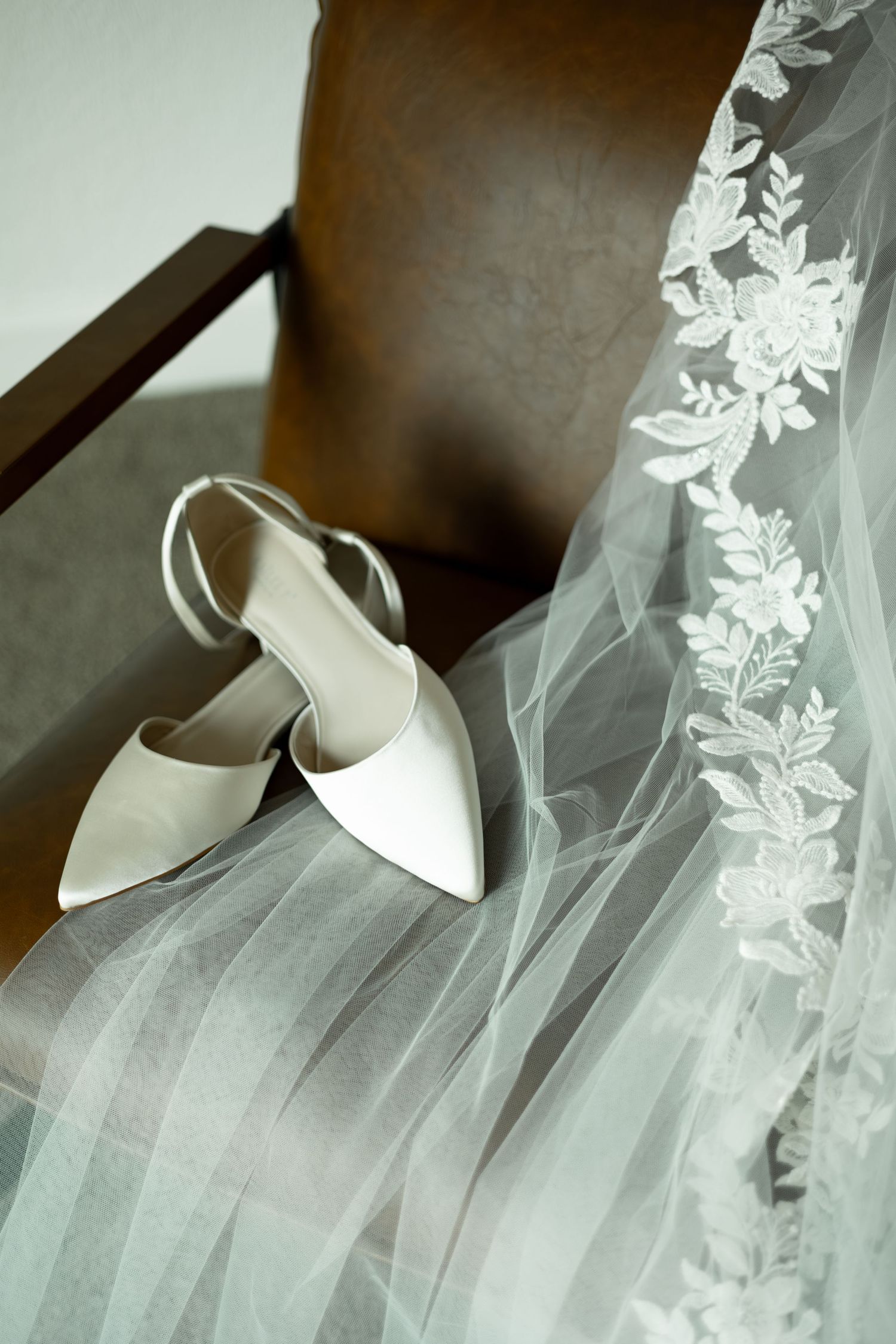 Elegant white bridal shoes rest beside delicate lace-trimmed wedding veil in soft lighting.