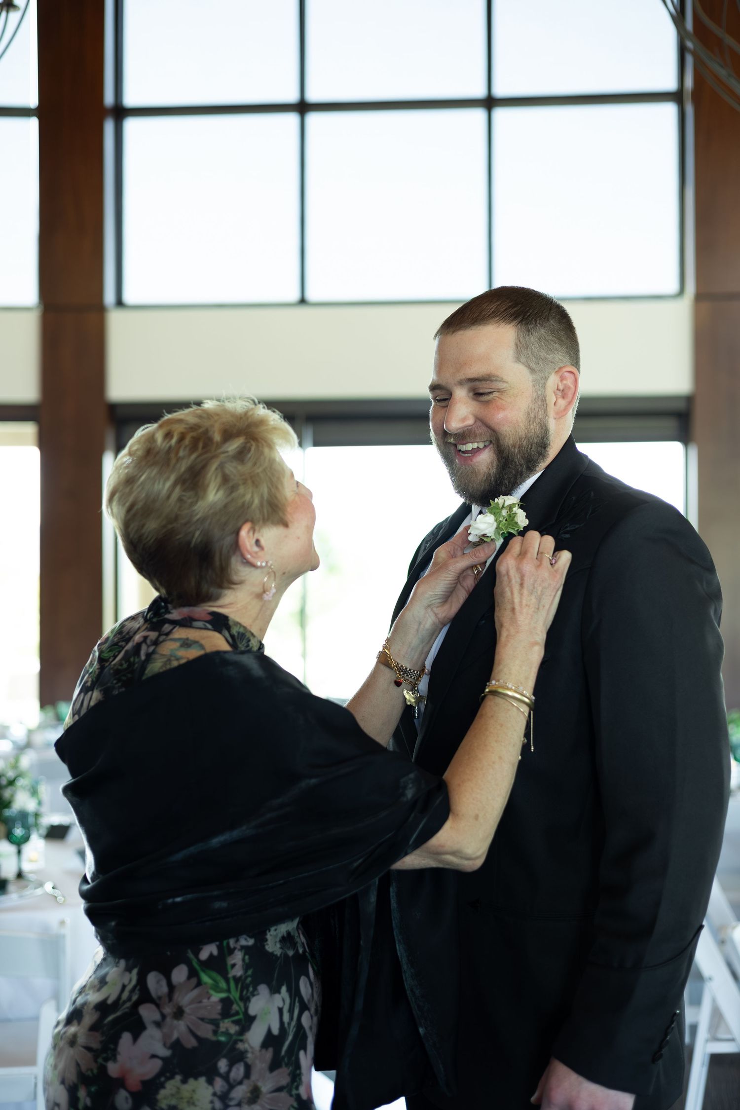 An older woman pins a boutonniere on a smiling bearded groom in a black suit at a wedding venue.