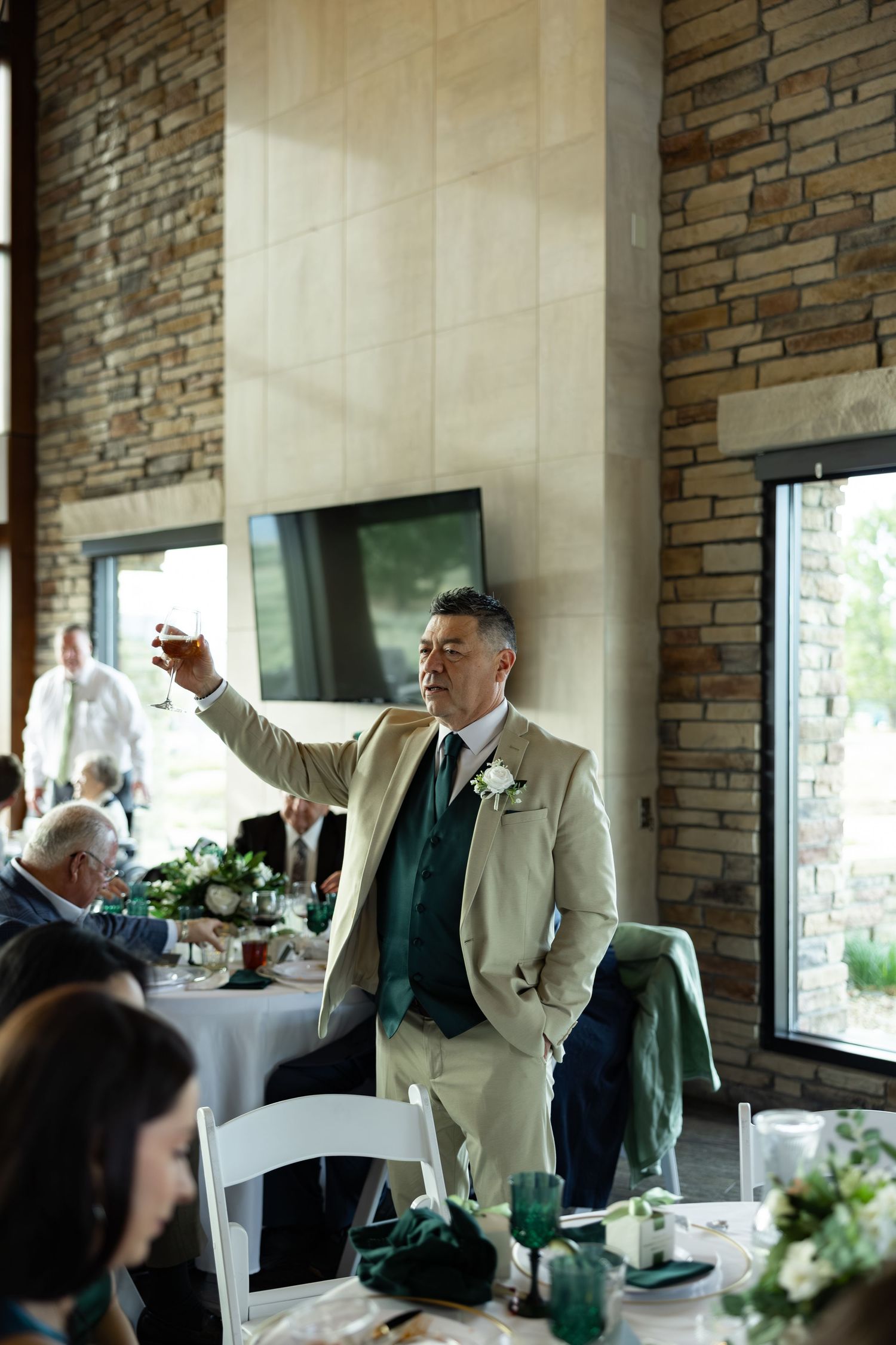 A man in a tan suit raises a glass for a toast at an elegant wedding reception with stone walls and floral decor.