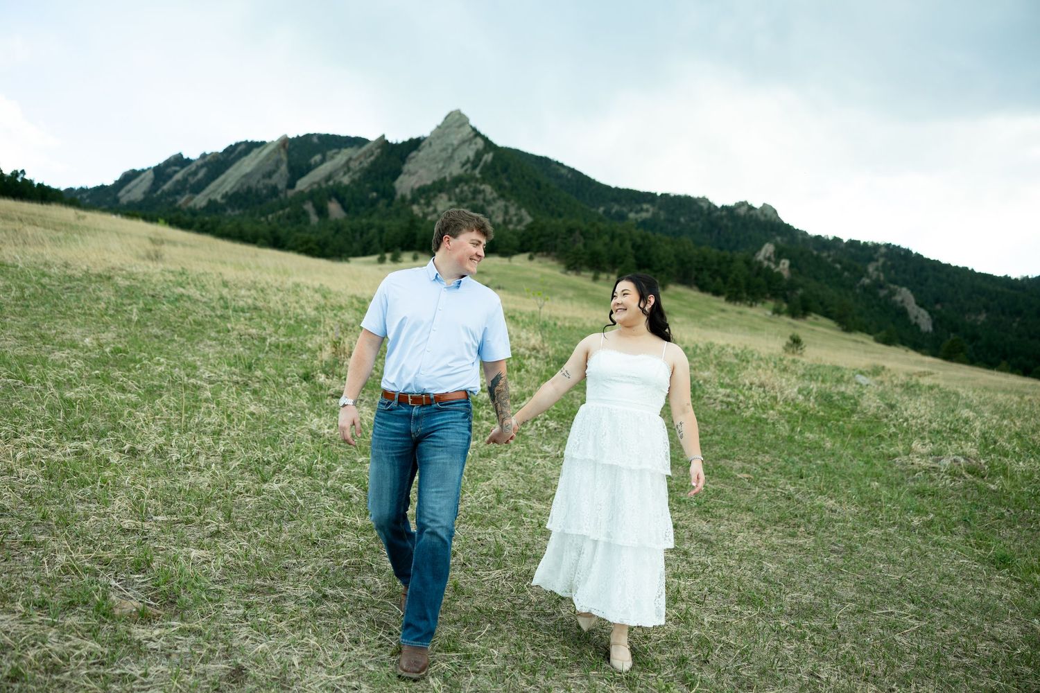 Couple holding hands walking through green meadow with Flatirons rock formations in Boulder Colorado background.