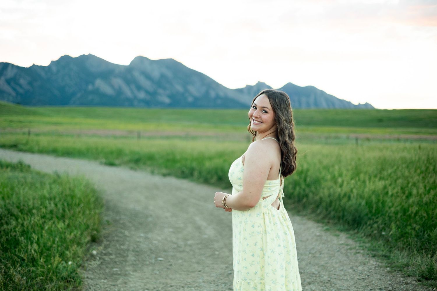 A figure in a flowing white dress poses in a verdant field with majestic mountains in the background during sunset.