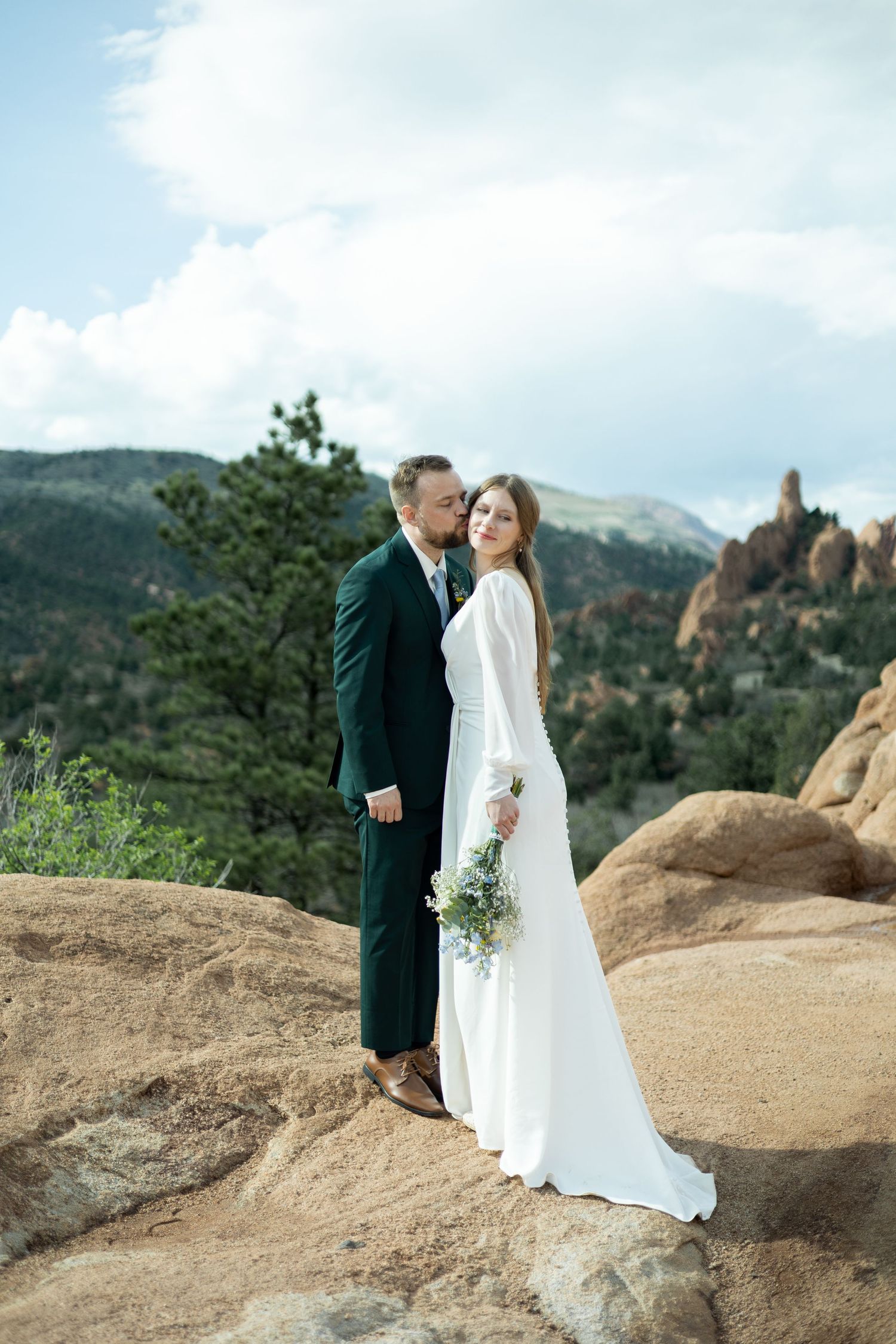 Bride and groom pose on rocky mountain terrain, groom kissing bride's cheek as she holds a wildflower bouquet.