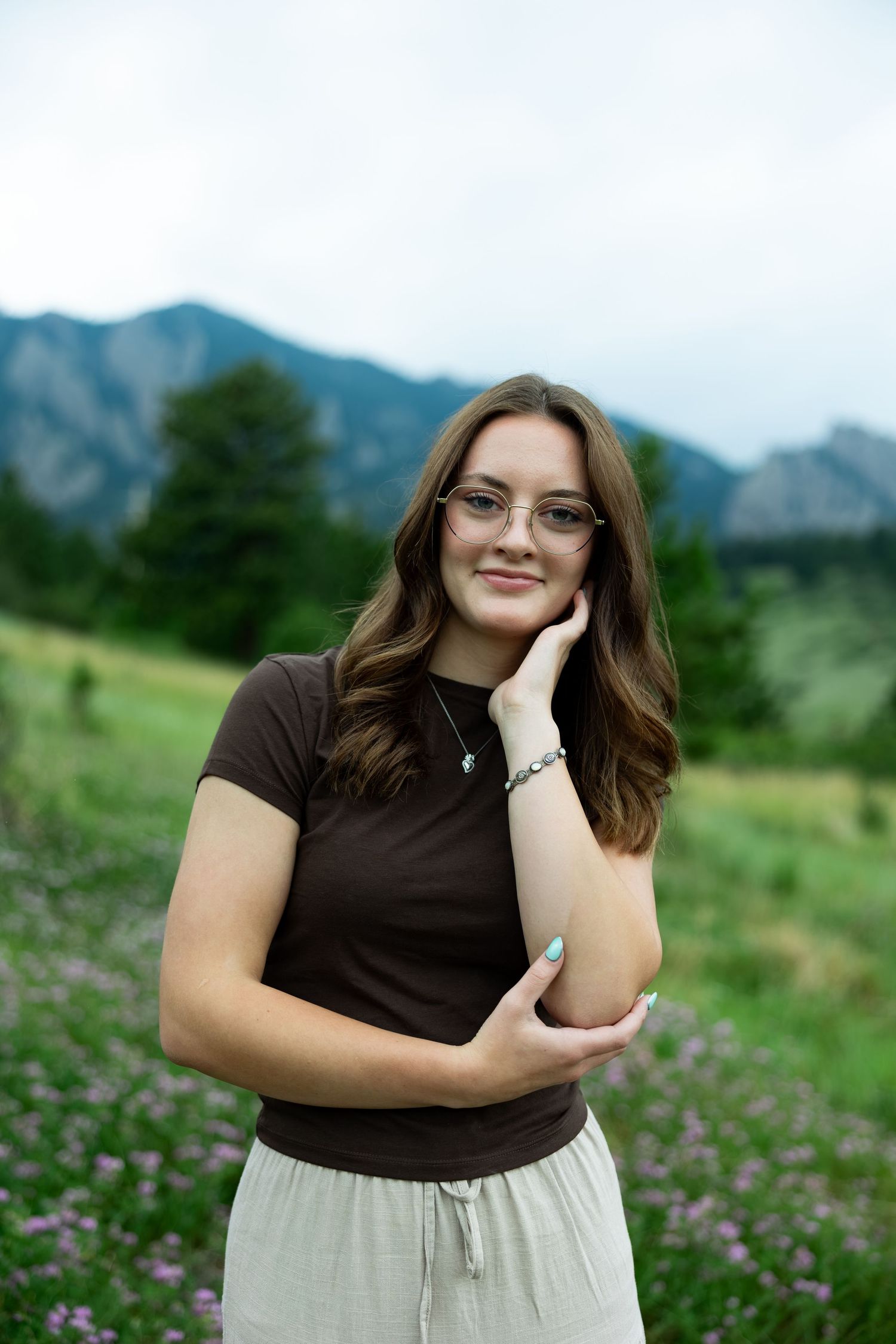 Young woman with glasses and brown hair poses in a mountain meadow with wildflowers and rocky peaks behind her.