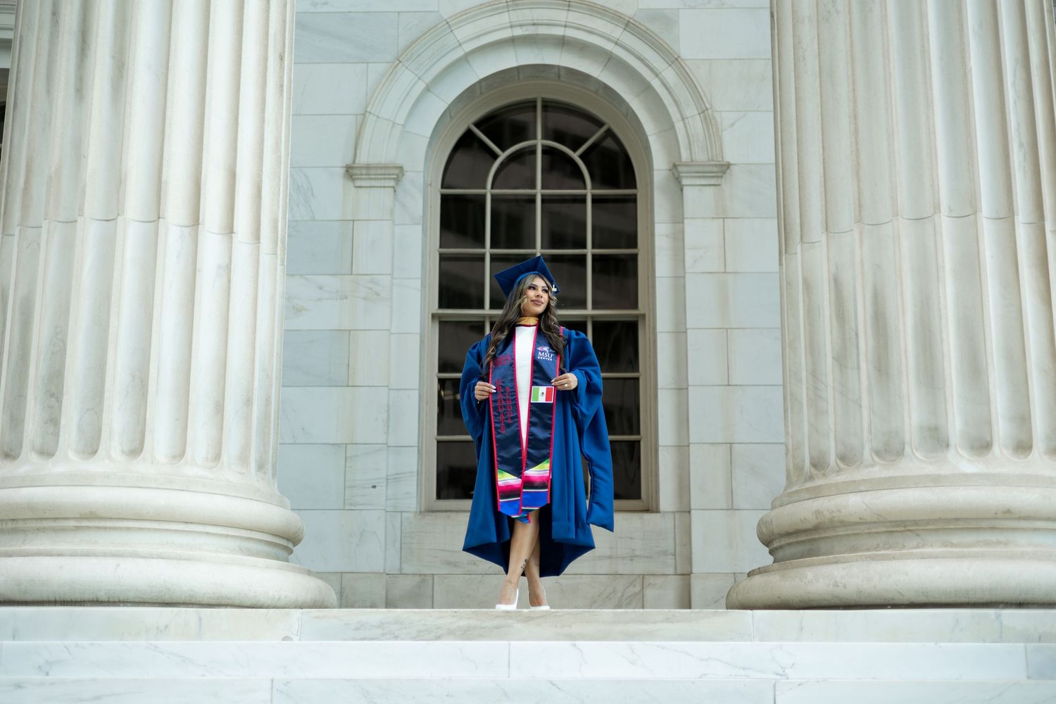 Graduates in traditional academic regalia pose in sequence between white stone columns of a classical building.