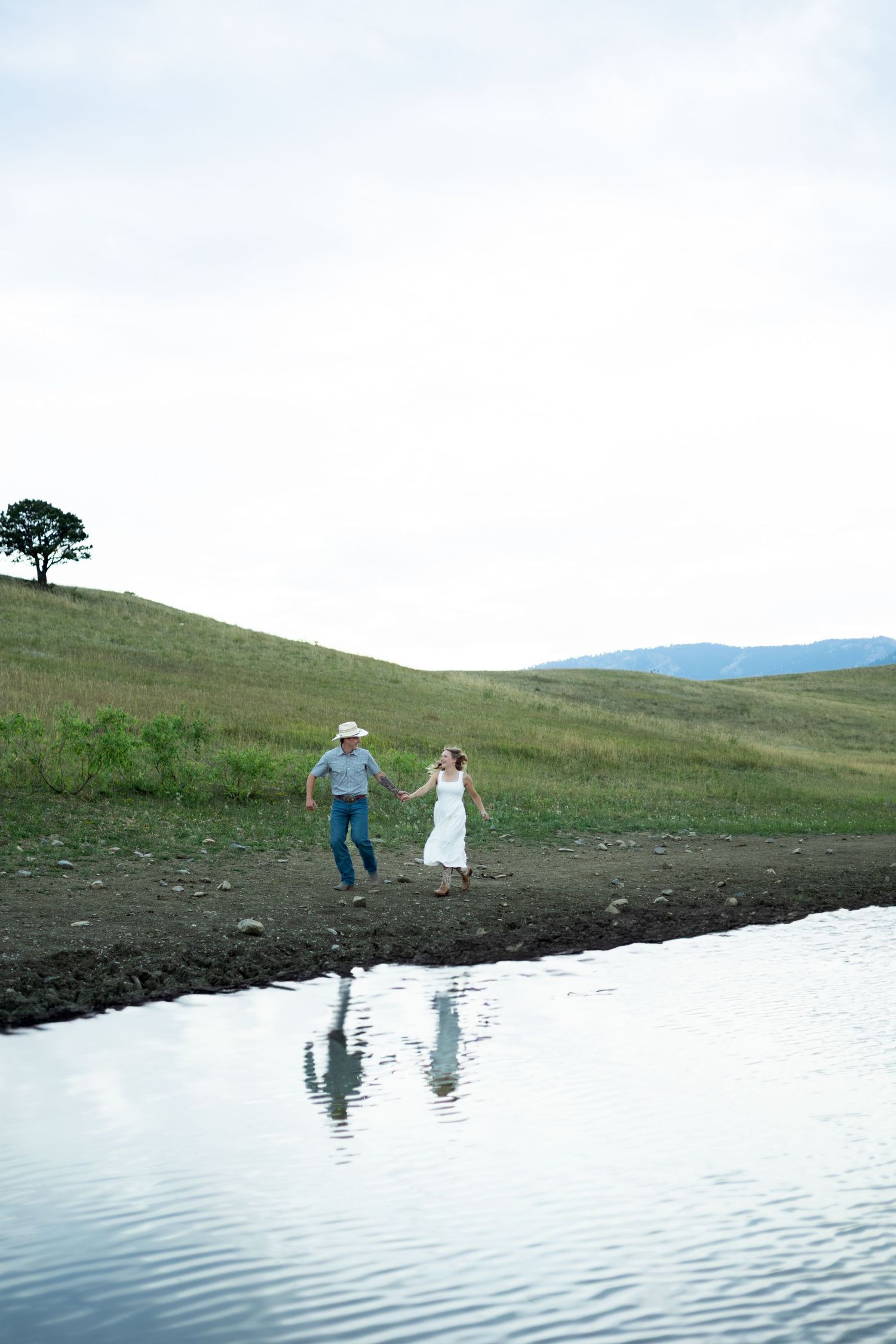 Two people in white outfits play and run along the edge of a calm lake surrounded by green rolling hills.