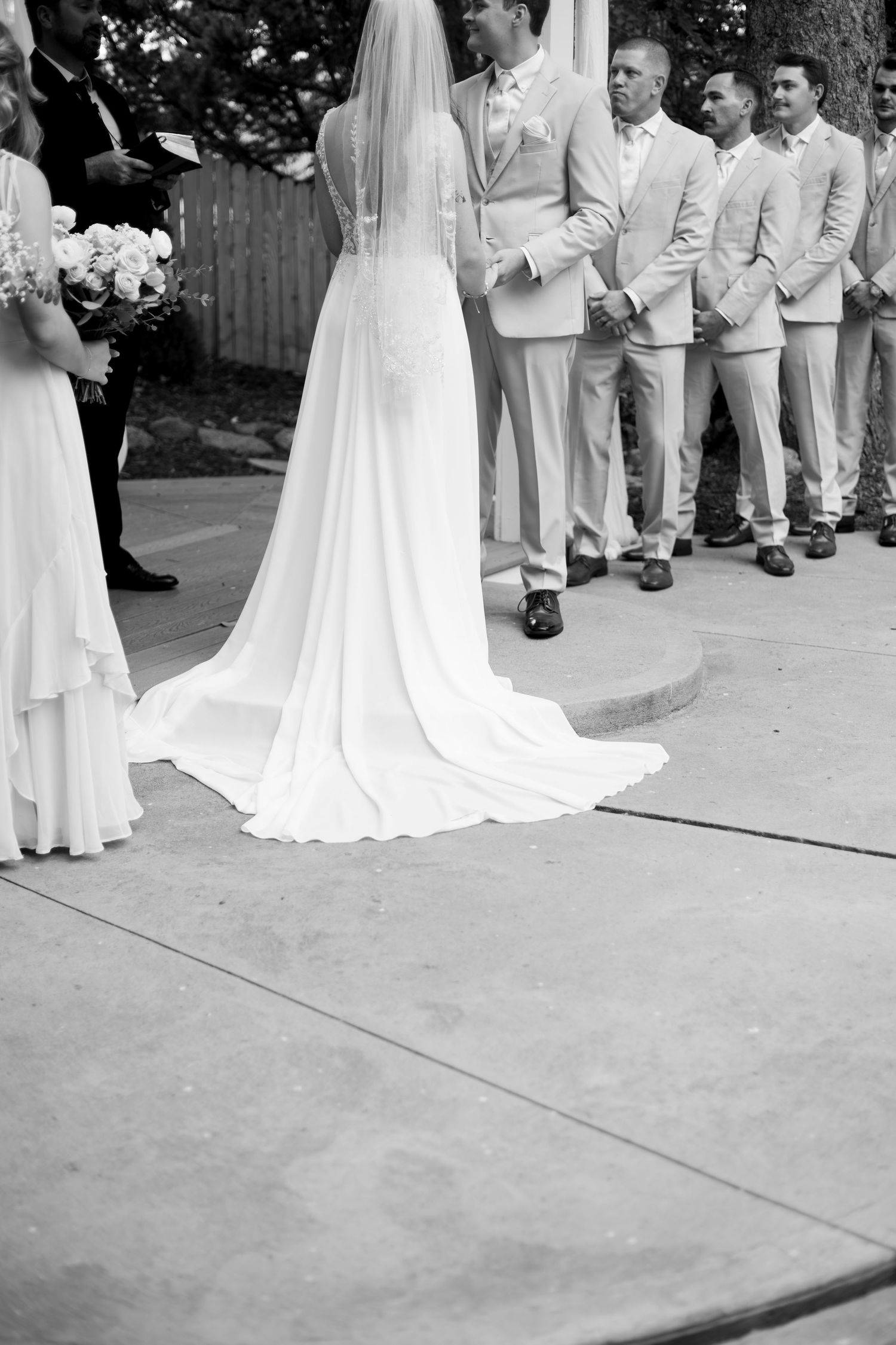 Black and white photo of a wedding ceremony gathered on a concrete patio with trees in the background.