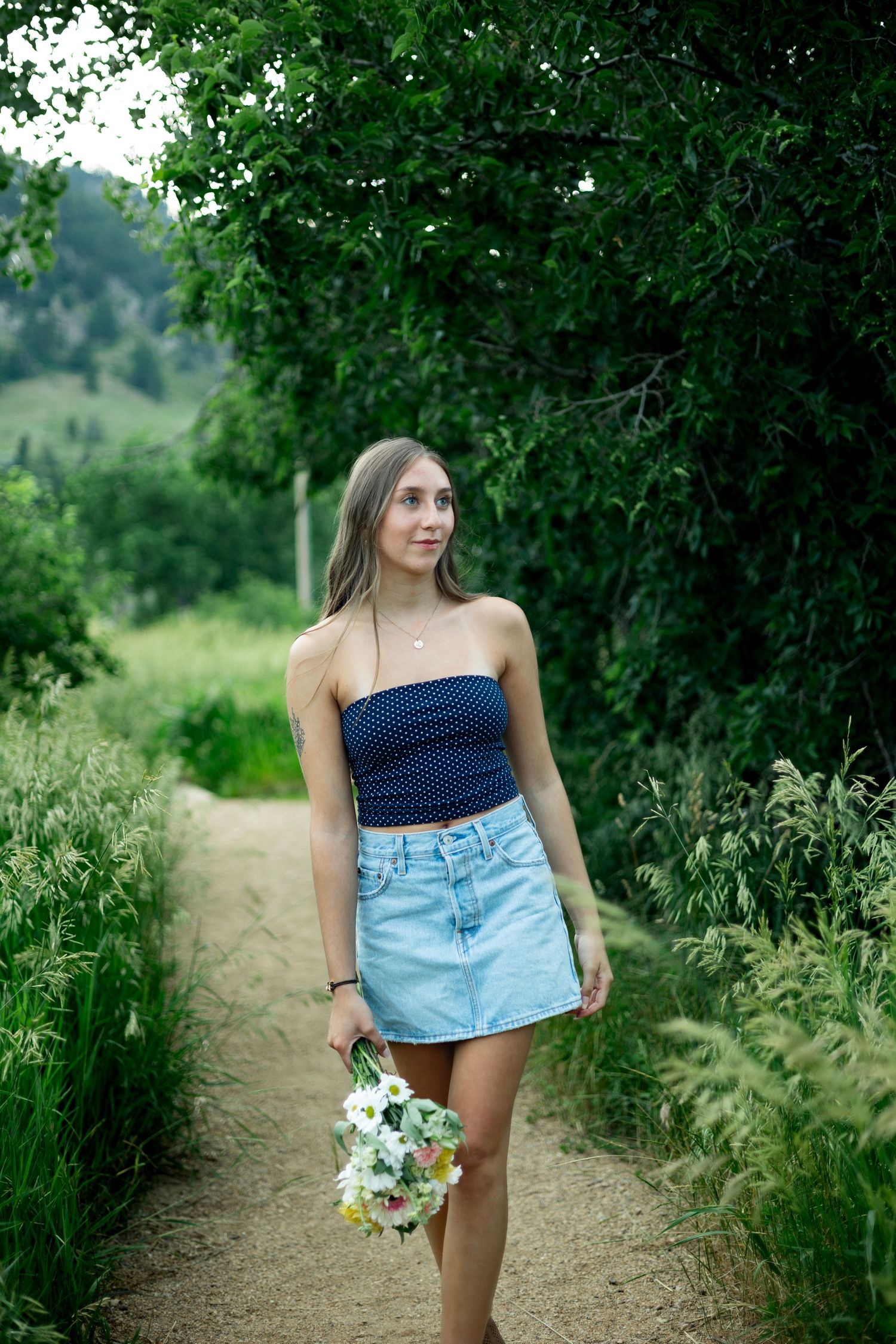 A woman in a navy polka dot top and denim skirt walks along a dirt path holding wildflowers on a summer day.