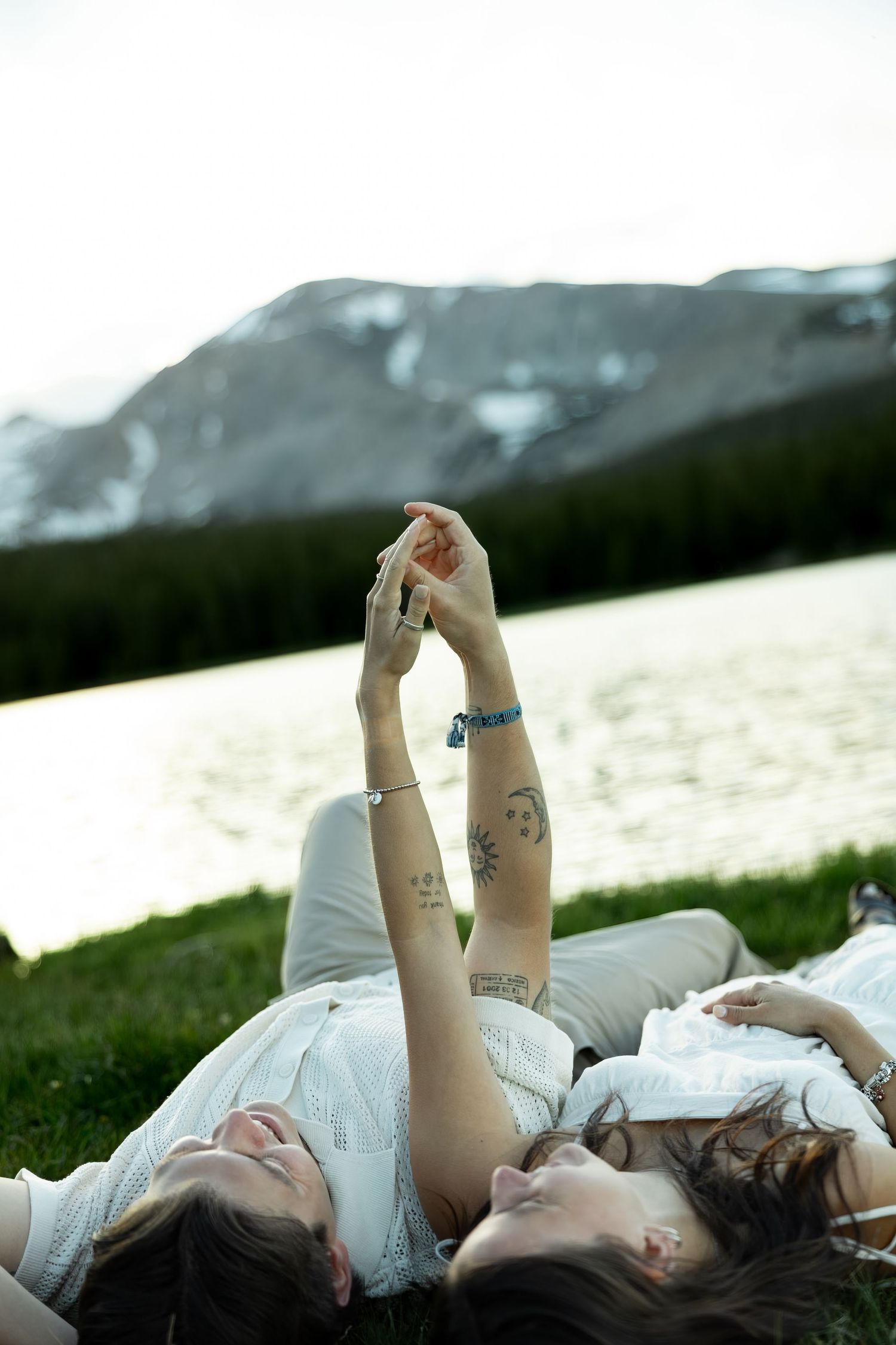 Two women lying on grass holding hands up, with a snow-capped mountain and lake in the background.