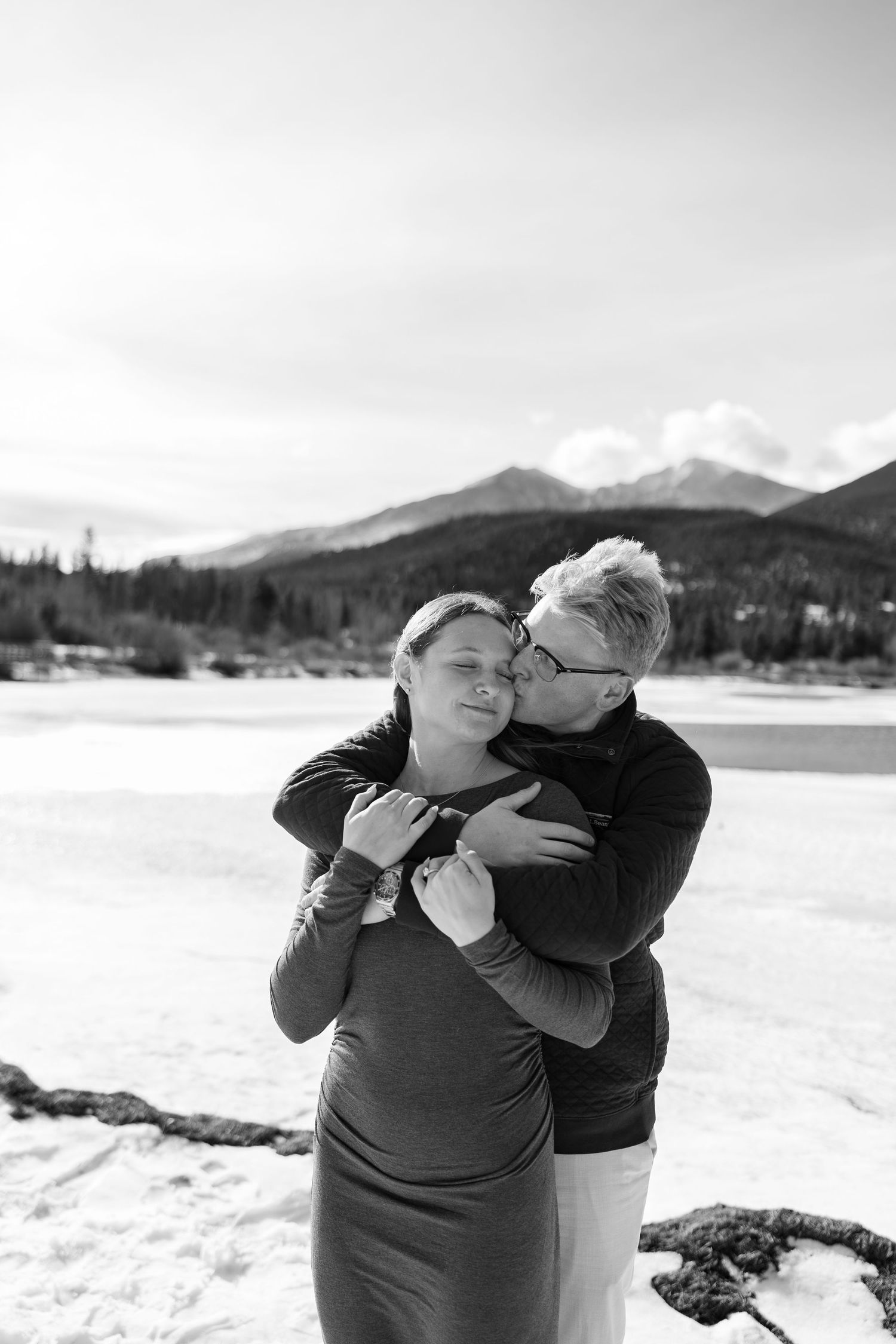 Couple sharing an intimate moment on a snowy landscape with mountains and a frozen lake behind them.