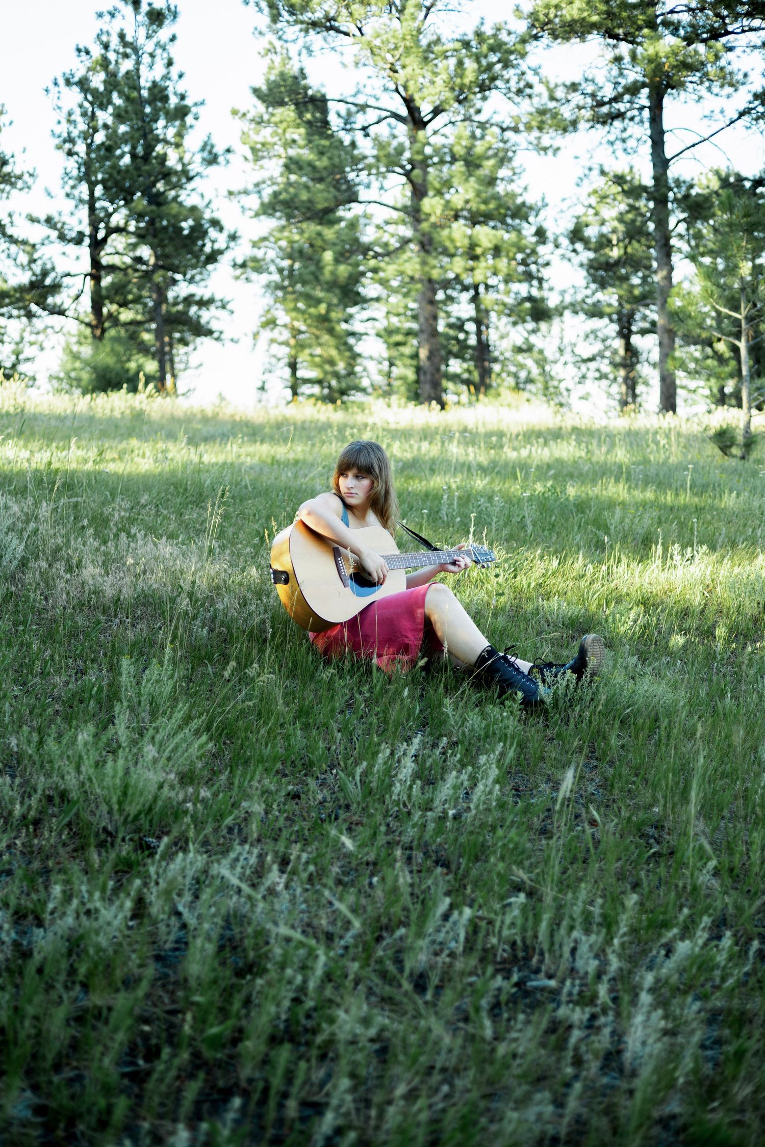 Young woman sitting in a green meadow playing acoustic guitar, surrounded by tall pine trees on a sunny day.