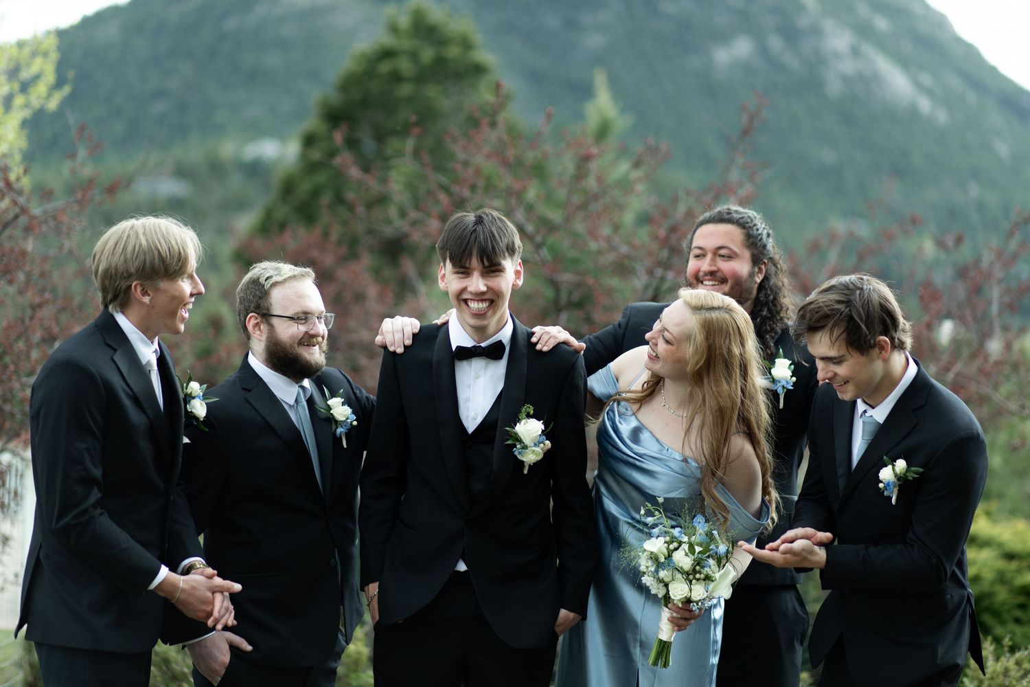 Group of formally dressed people laugh together against scenic mountain backdrop.