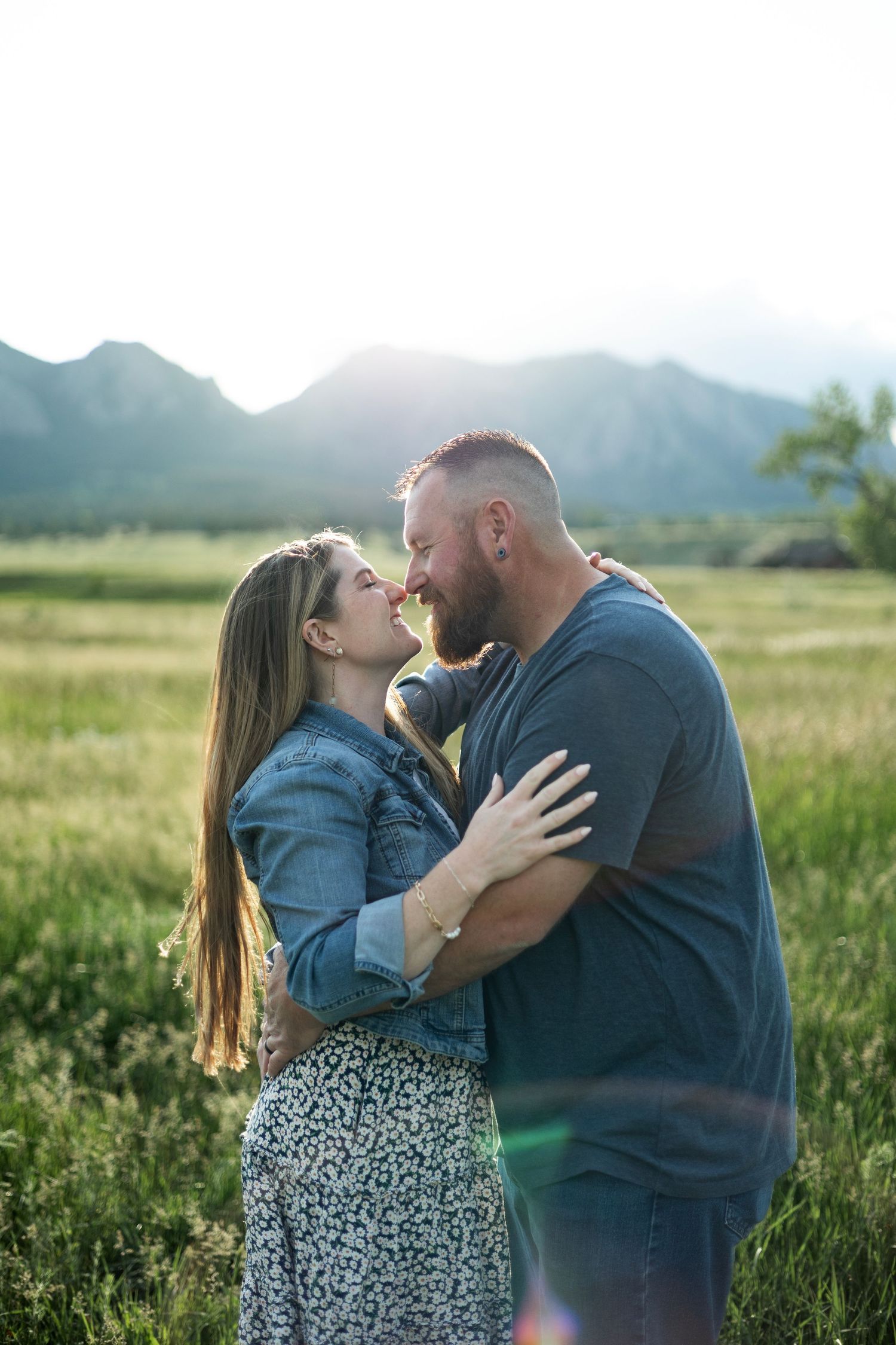Couple embracing and smiling at each other in a sunlit green field with mountain backdrop during golden hour.