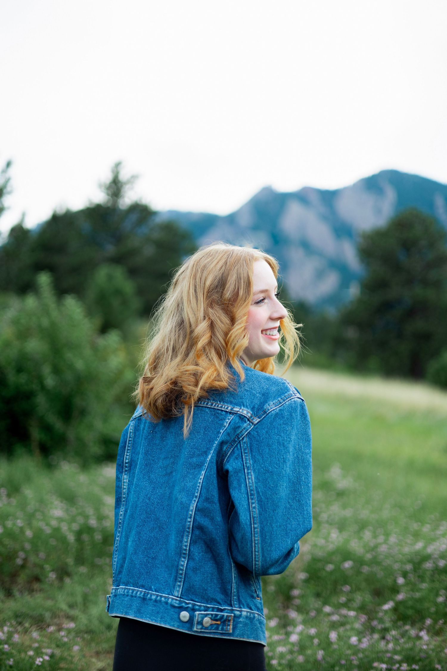 Young woman with red hair in denim jacket smiling outdoors with mountain and green meadow backdrop.
