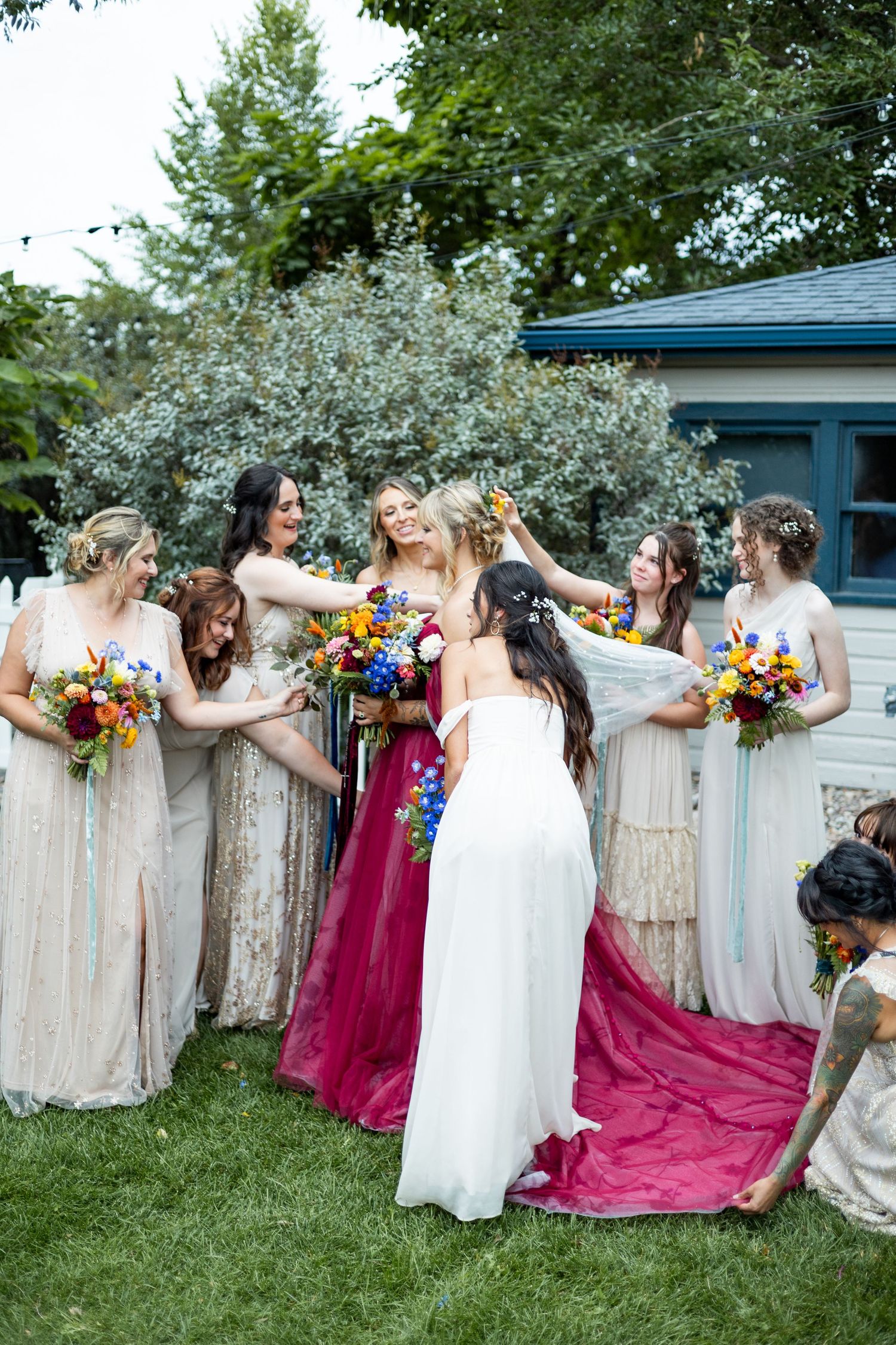 Bridesmaids in light dresses gather around two people in white and burgundy gowns with bouquets.