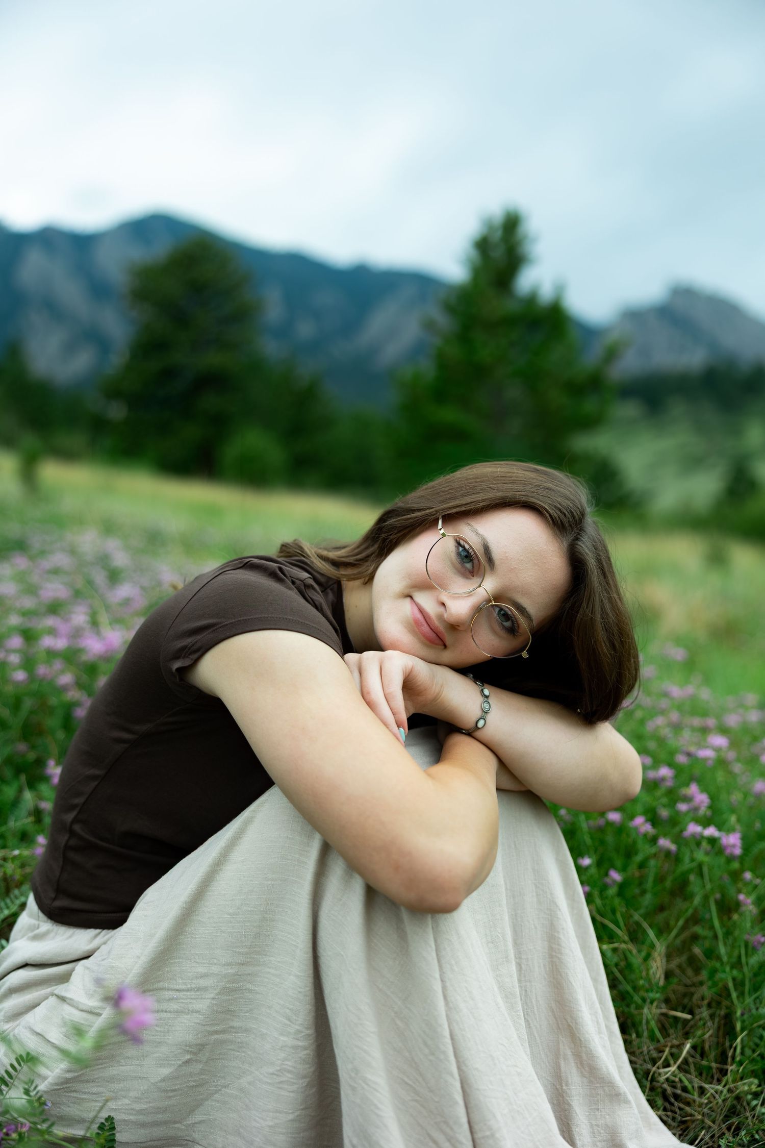 Young woman with glasses resting on knees in a wildflower meadow with mountains and trees in background.