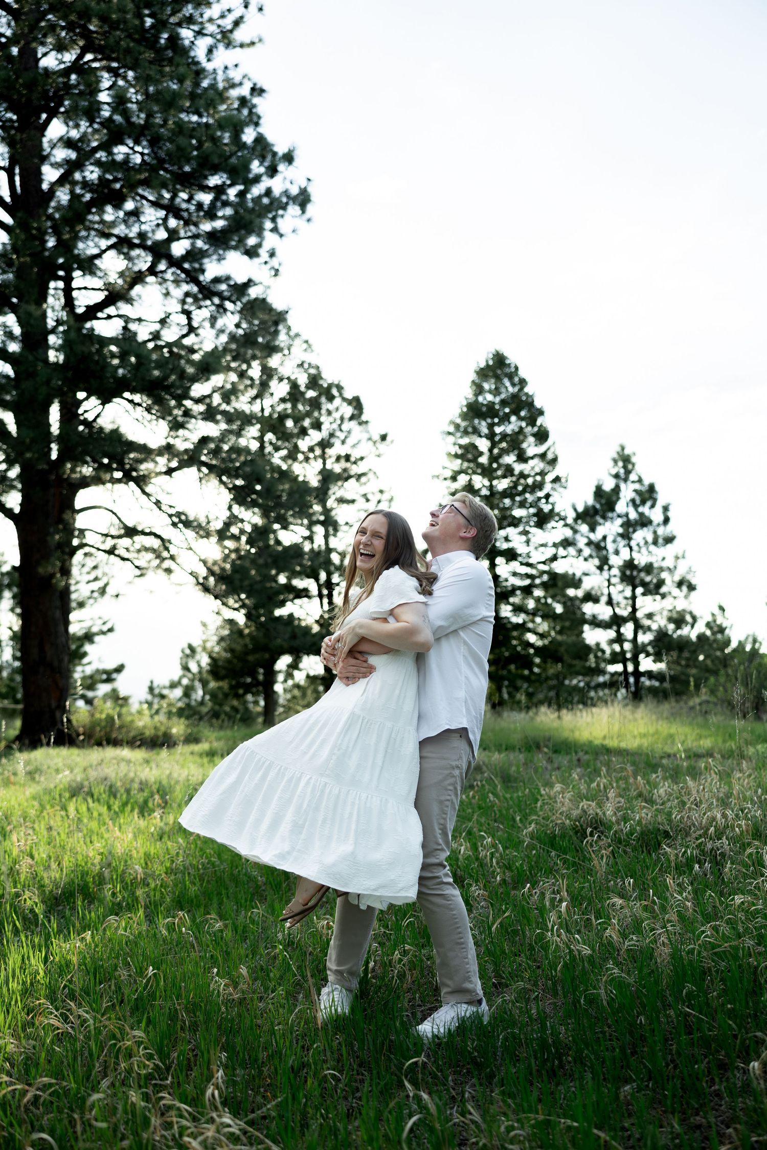 Happy couple dancing and laughing together in a lush green meadow surrounded by tall pine trees.