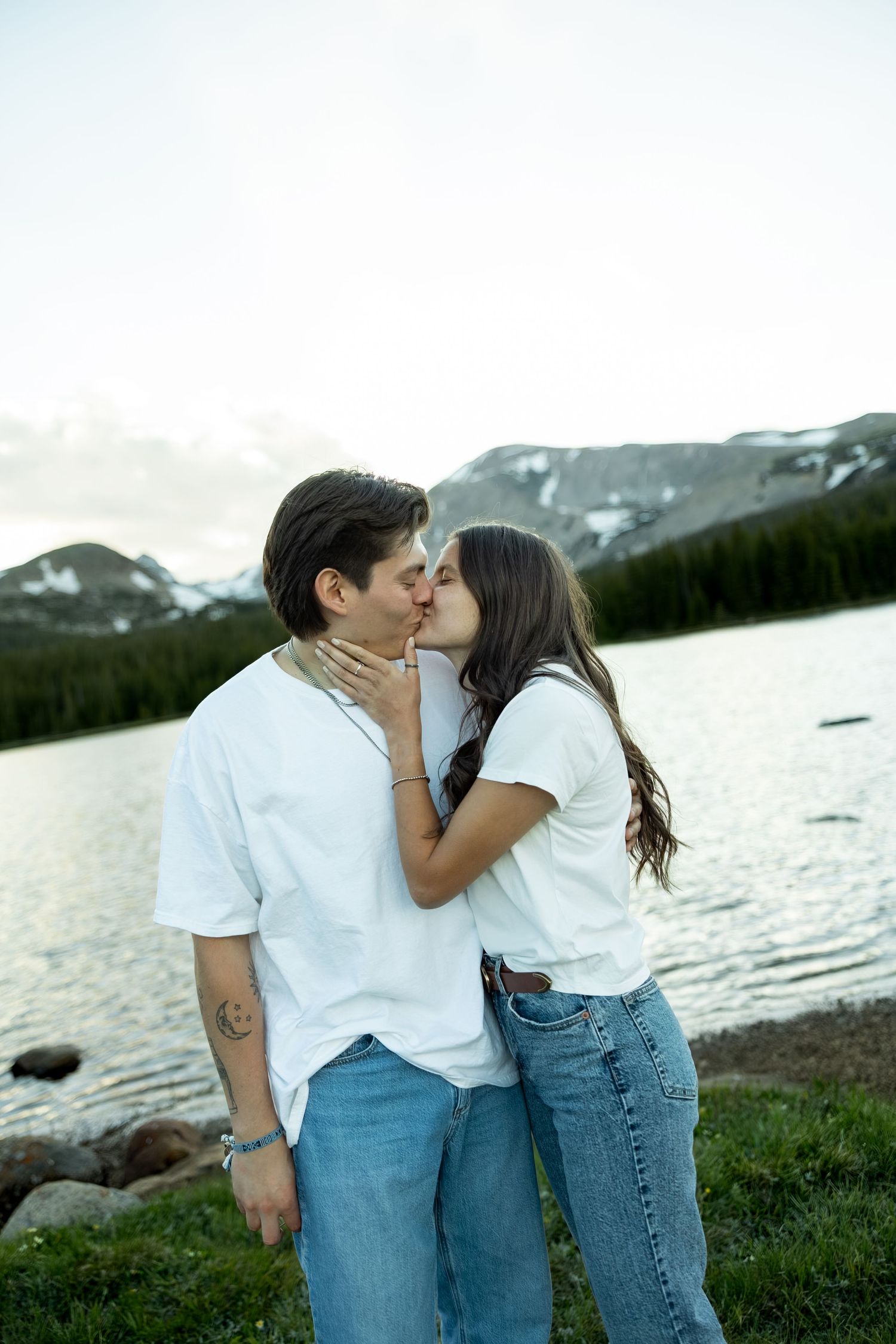 Young couple sharing a kiss by a mountain lake, both wearing white tops and jeans in scenic nature setting.