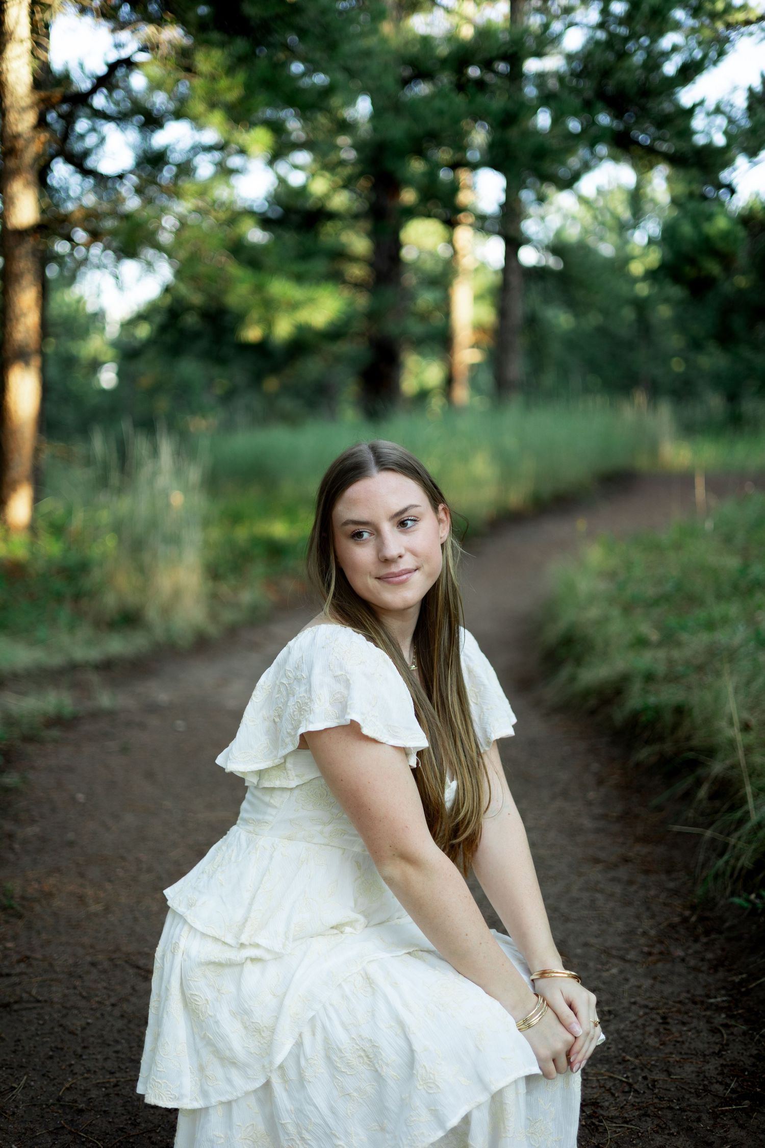 A peaceful forest scene features someone in a white dress sitting on a dirt path surrounded by towering pine trees.