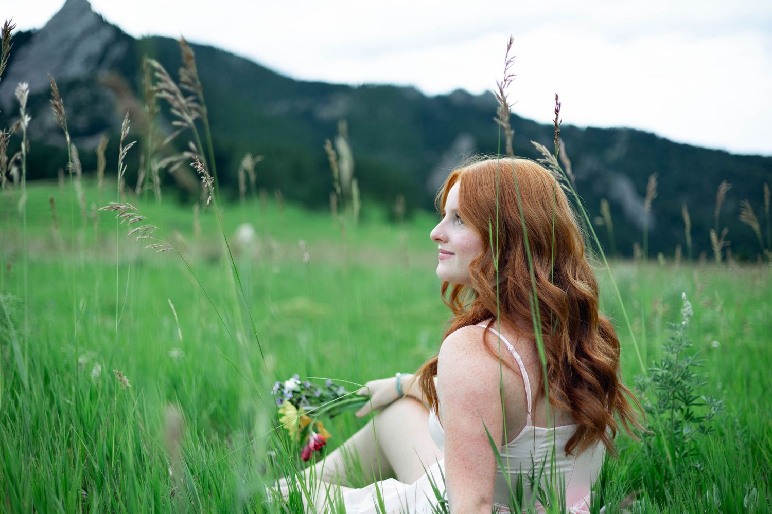 Woman with long red hair sits in a green summer meadow with tall grass blowing in the breeze.